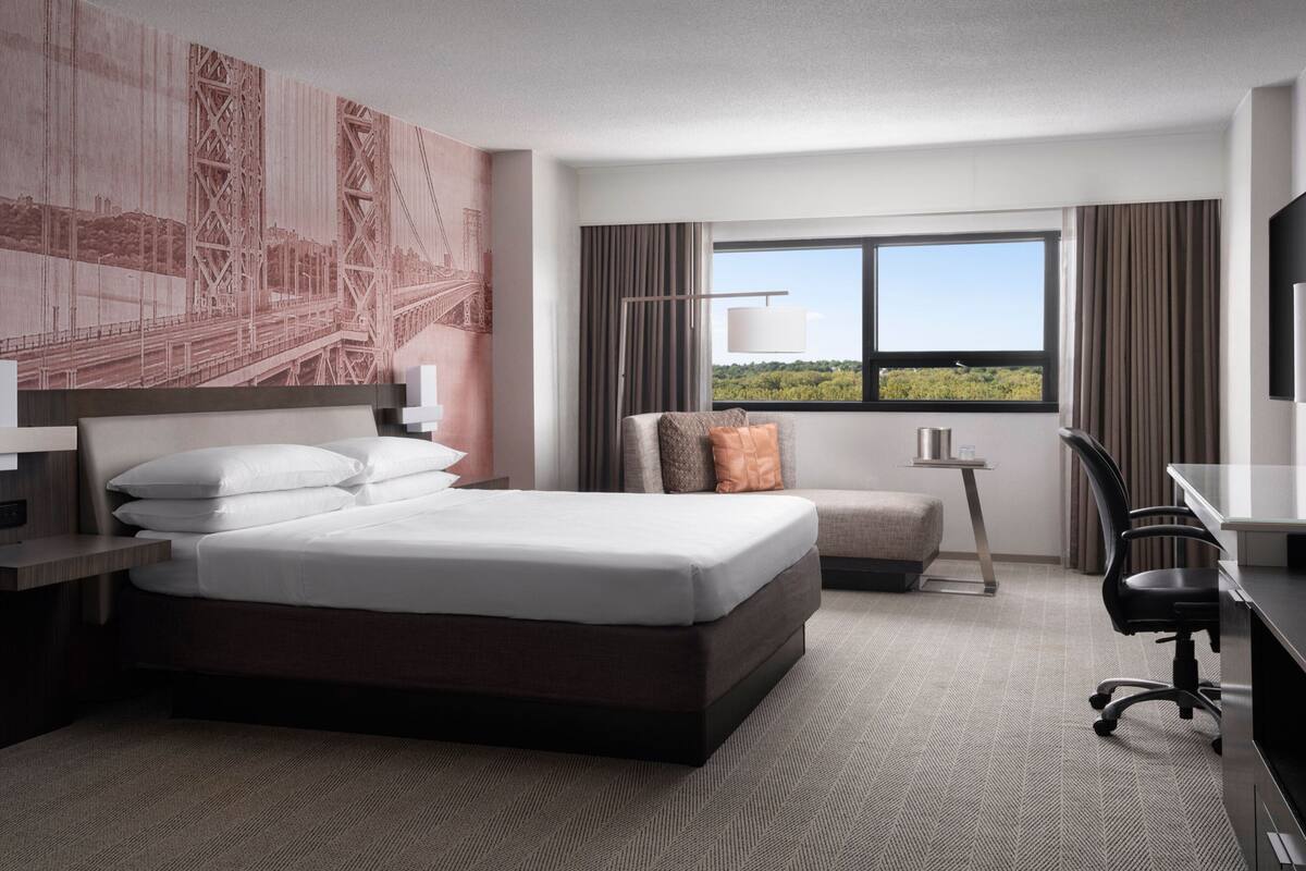 Ein Hotelzimmer mit einem großen Bett, einem Schreibtisch, einem Stuhl, einer Lampe und Blick auf Grün.