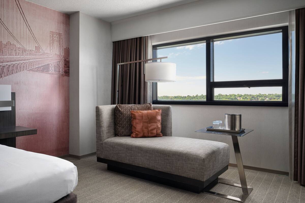 Ein Hotelzimmer mit Bett, einer Couch, einem Tisch mit Lampe, einem Fenster mit Aussicht und einer Wand mit Wandgemälde.