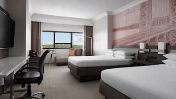 Une chambre d’hôtel équipée d’un bureau, de deux lits, d’une télévision et d’une grande fenêtre donnant sur un beau paysage.