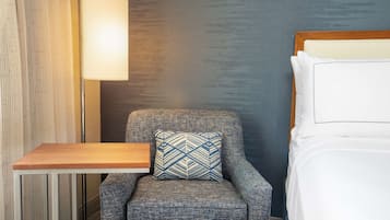 A hotel room with a grey armchair, a bedside table, a lamp, and a bed with white linens.