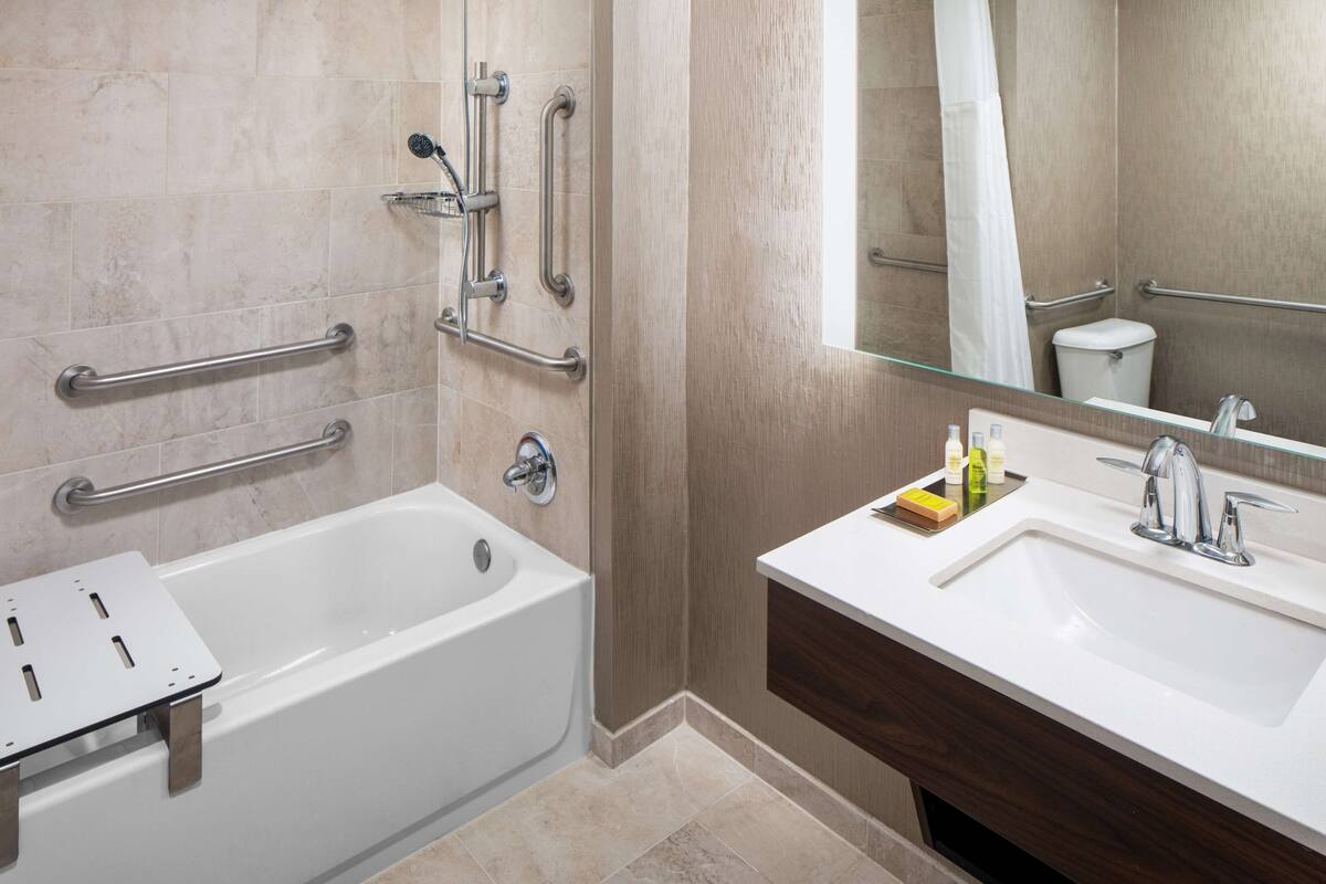 Une salle de bain moderne avec une baignoire, un meuble-lavabo et un miroir. Des barres d’appui sont fixées aux murs, et il y a une pomme de douche.