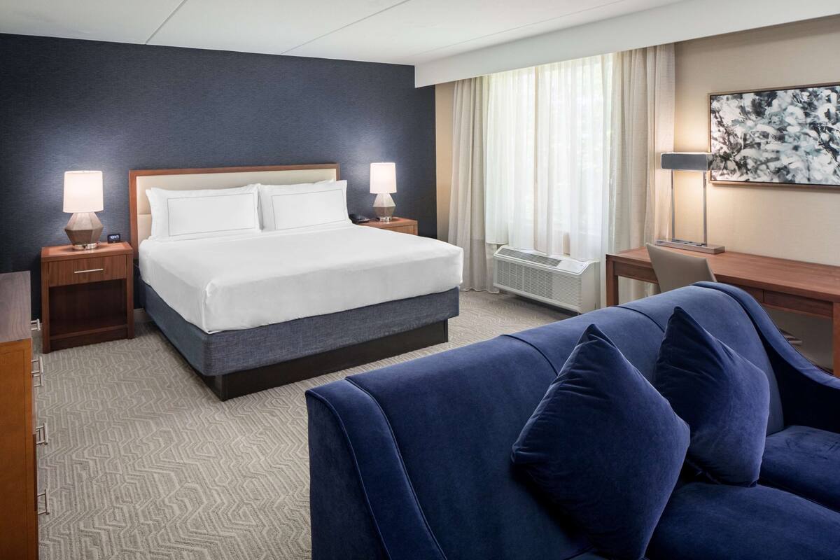 Une chambre d’hôtel avec un grand lit, deux tables de chevet avec des lampes, un bureau avec une chaise et une grande fenêtre avec des rideaux.