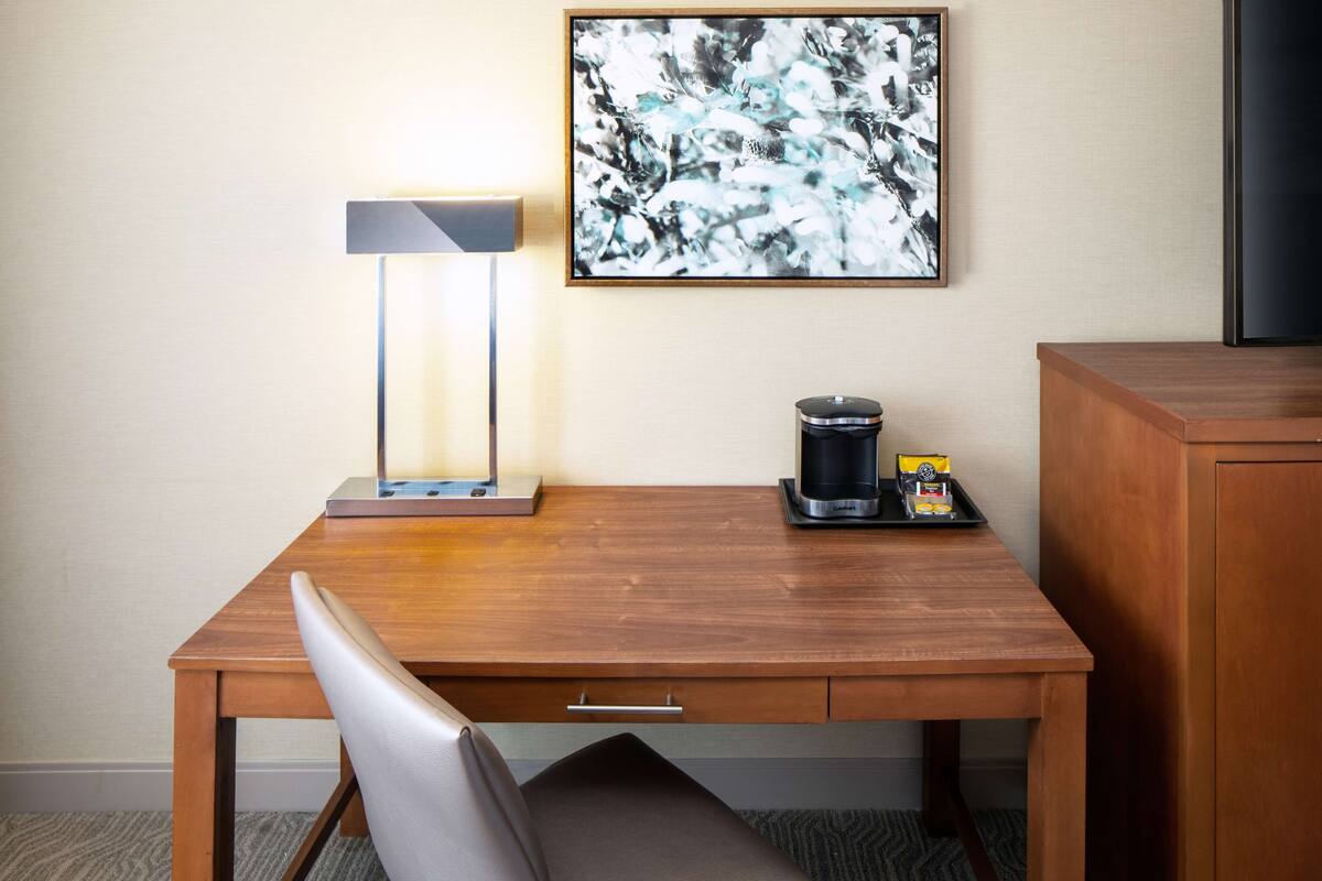 Une chambre d’hôtel avec un bureau en bois, une lampe moderne, une machine à café et un tableau abstrait encadré accroché au mur.