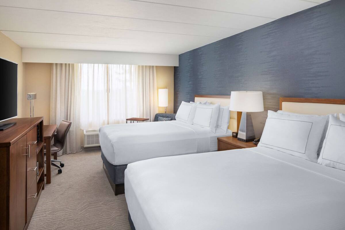 A hotel room with two beds, a desk, a TV, and a window with curtains.