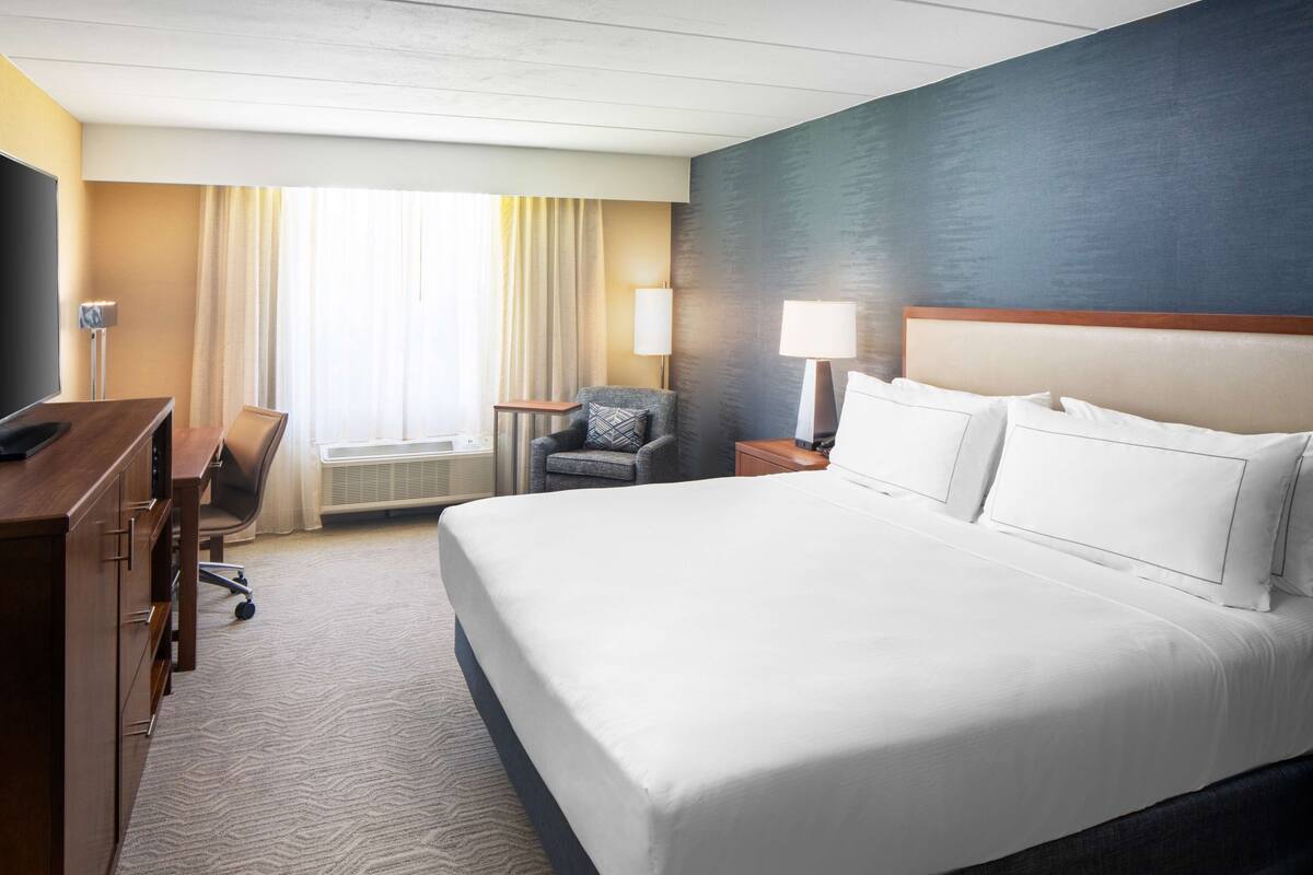 Une chambre d’hôtel avec un grand lit, un bureau, une chaise, une lampe et une fenêtre avec des rideaux.