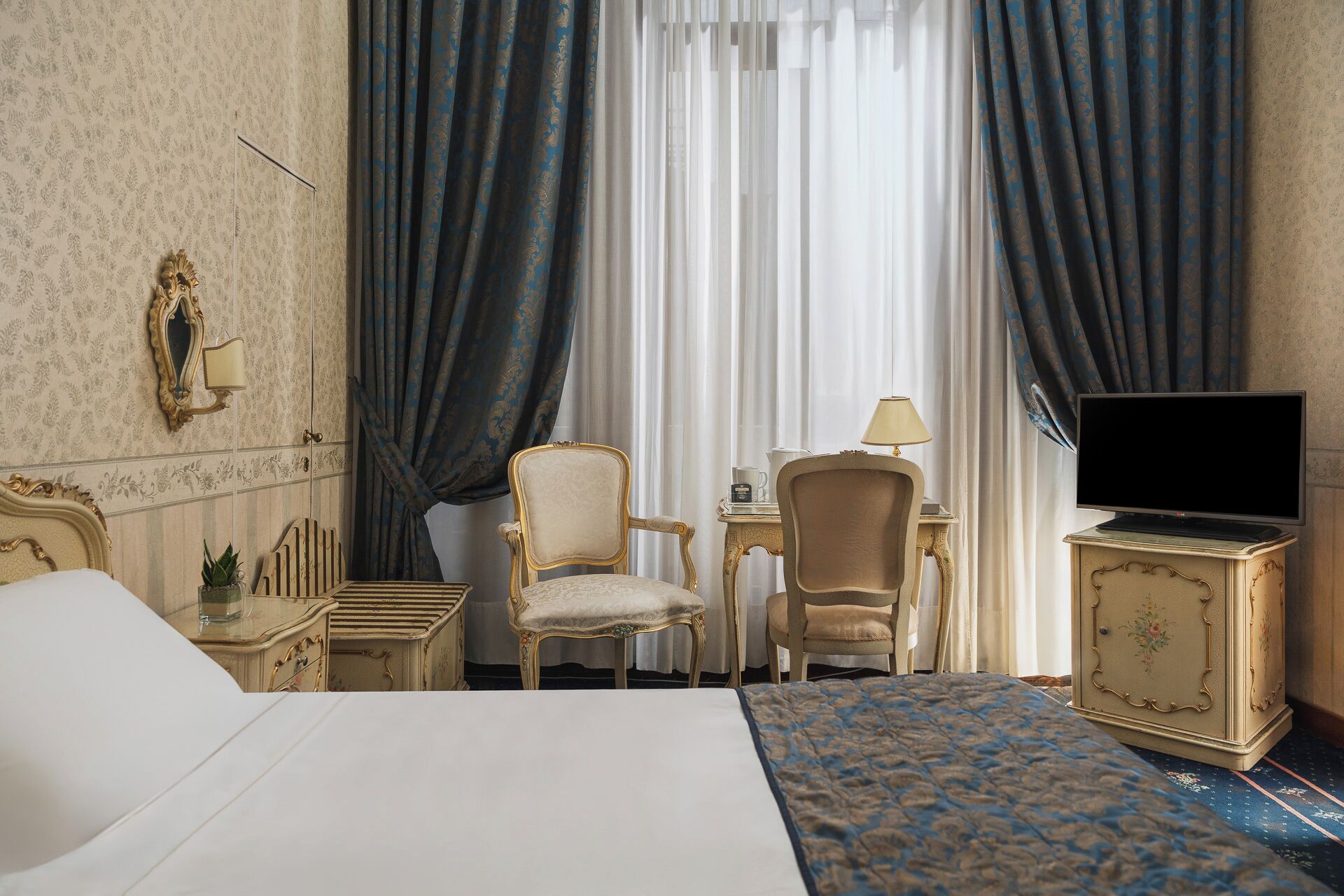 A hotel room with a bed, two chairs, a desk, a television, and curtains.
