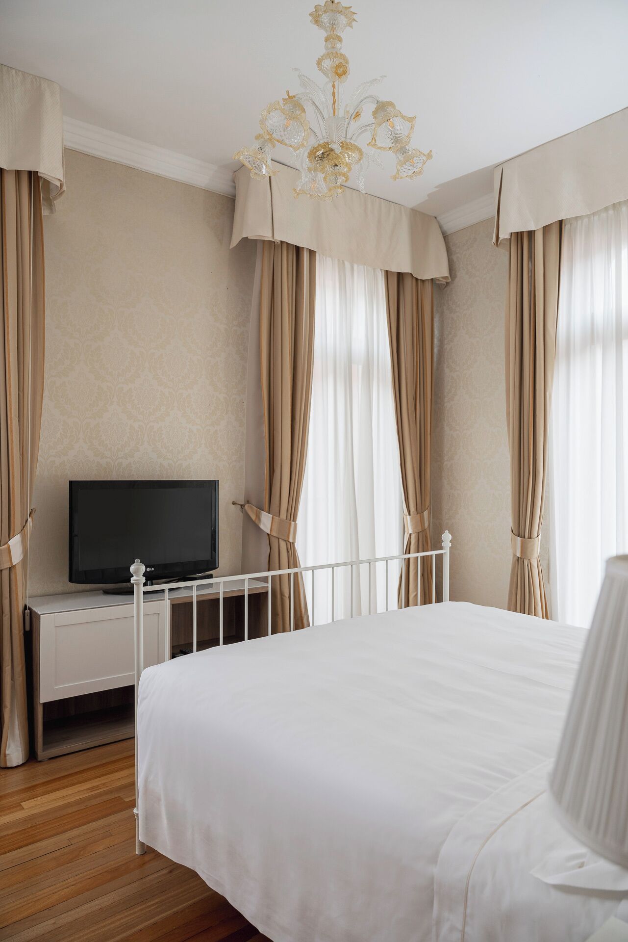 A hotel room with a large bed, a TV on a white cabinet, beige curtains, and a chandelier.