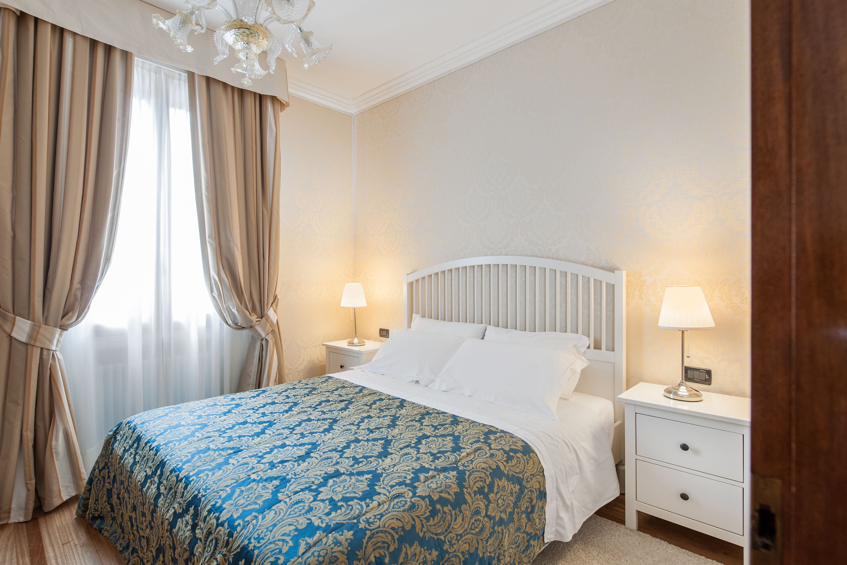 A bedroom with a bed, two bedside tables with lamps, a window with curtains, and a chandelier.