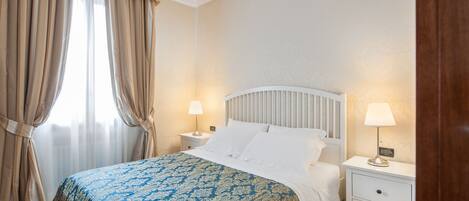 A bedroom with a bed, two bedside tables with lamps, a window with curtains, and a chandelier.