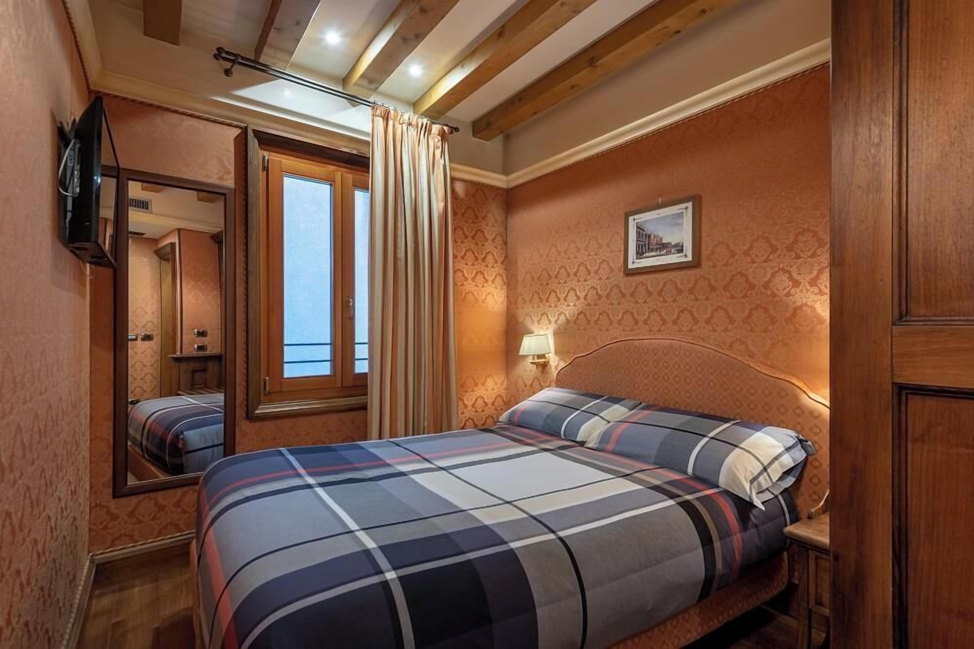A bedroom with a bed, a mirror, a window with curtains, a wall-mounted TV, and a framed picture.
