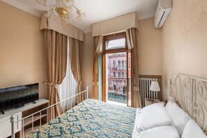 A hotel room with a bed, a TV, a nightstand, and a view of a building through the window. - Hotel Montecarlo (Venice)