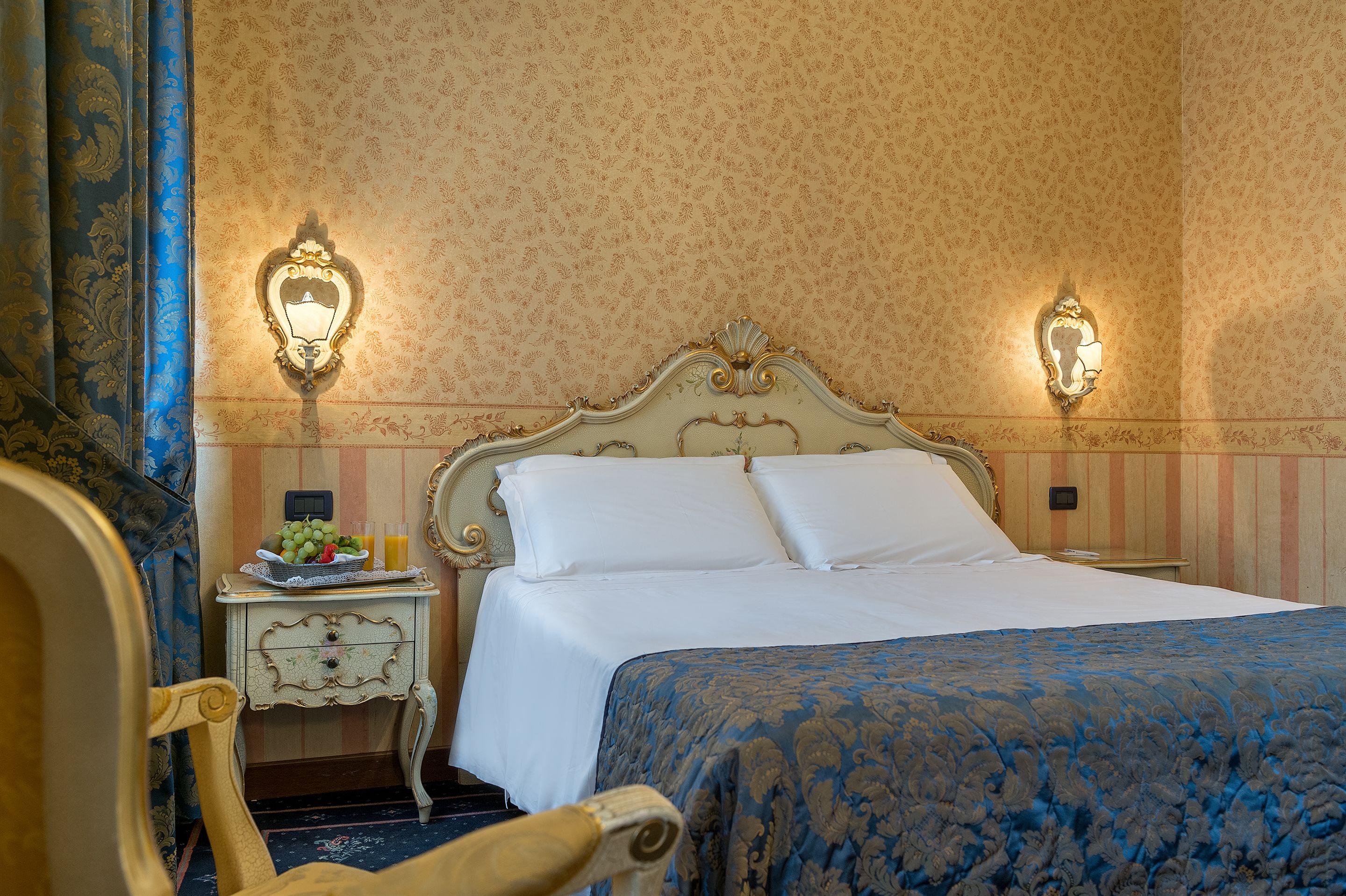 A hotel room with a bed, a nightstand with a fruit bowl and a lamp, and a chair.