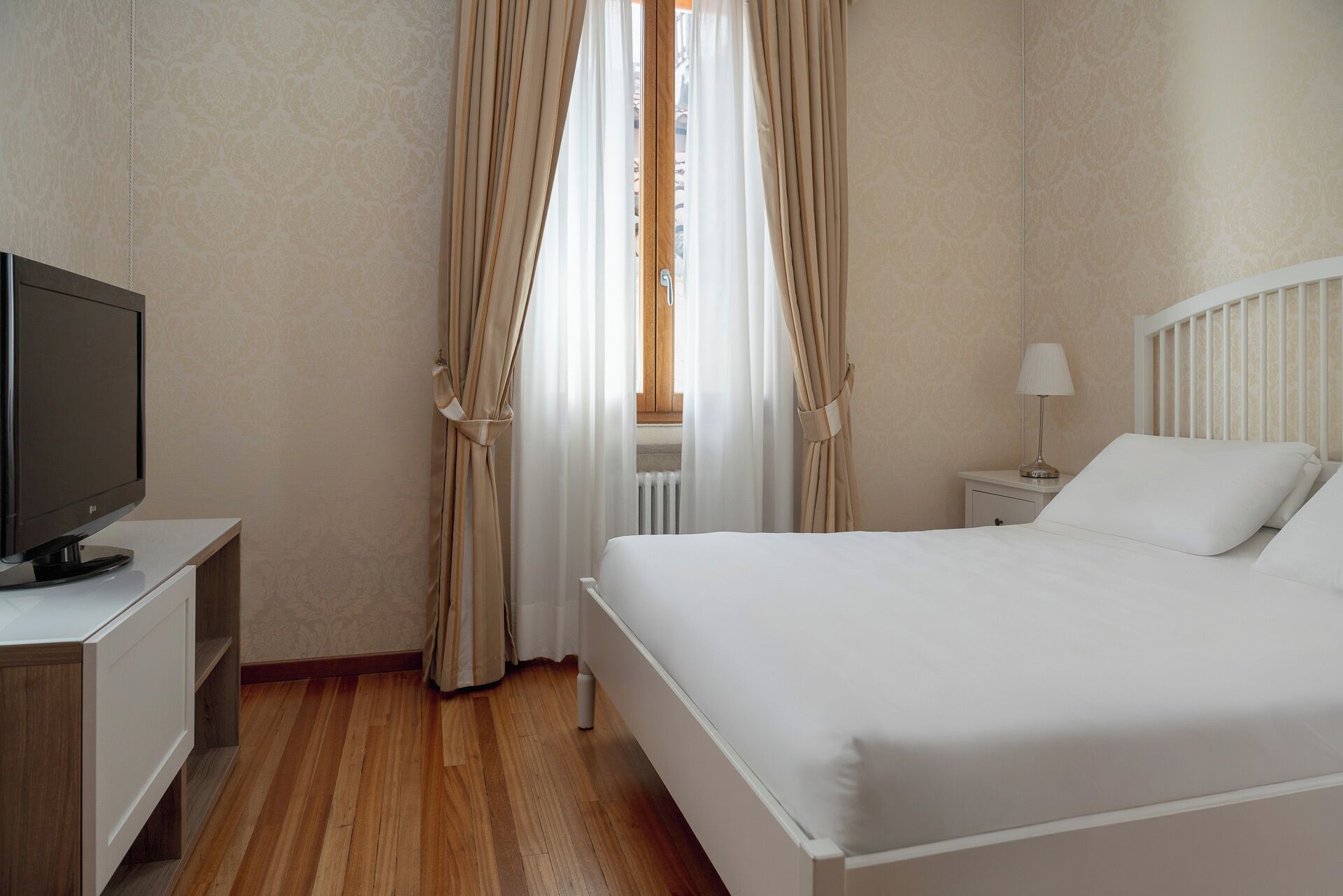 Habitación de hotel con cama, mesita de noche con lámpara, televisor sobre un mueble bajo y cortinas.