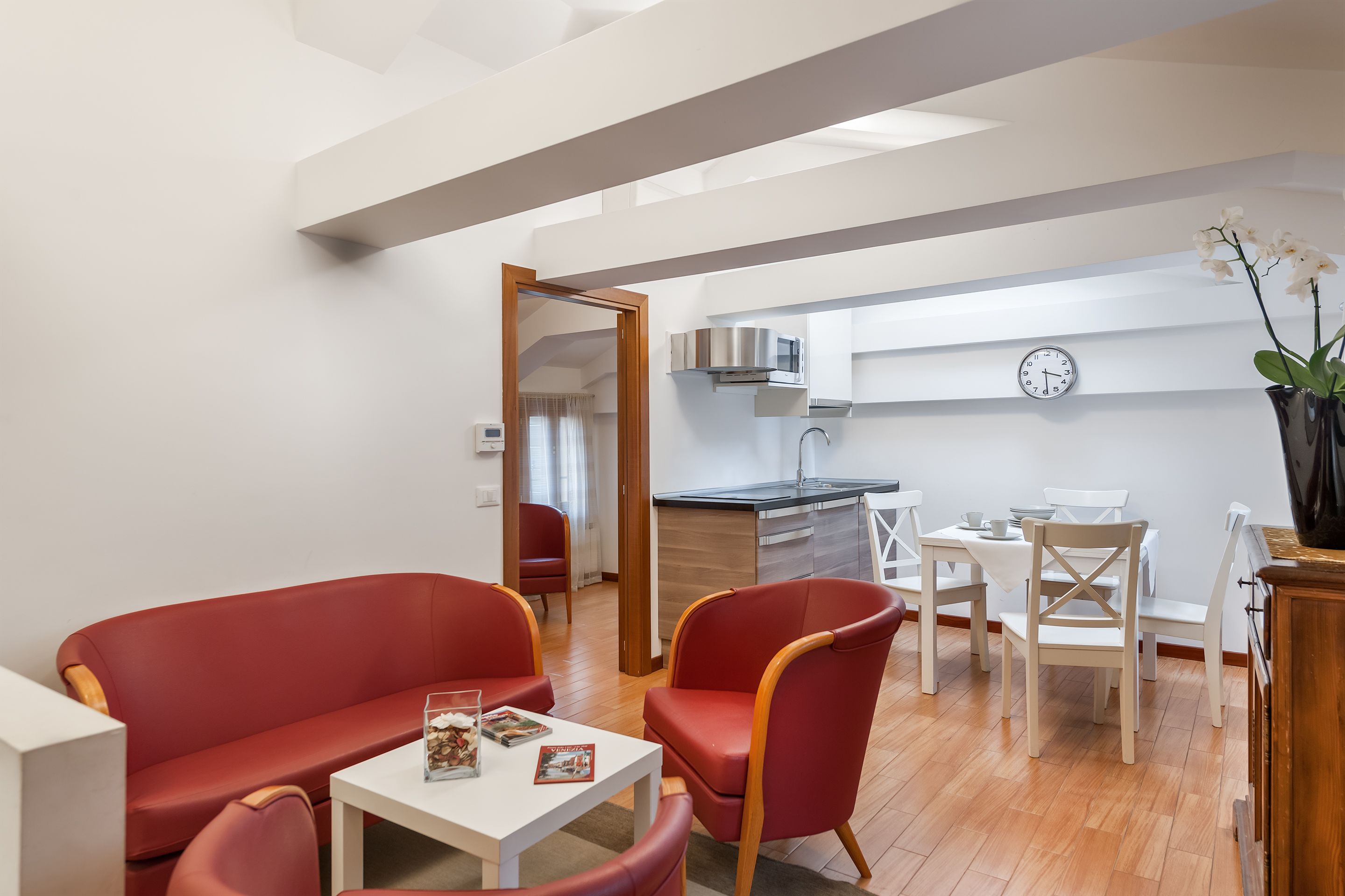 A modern living room with red furniture, a dining area with white chairs, and a kitchen in the background.