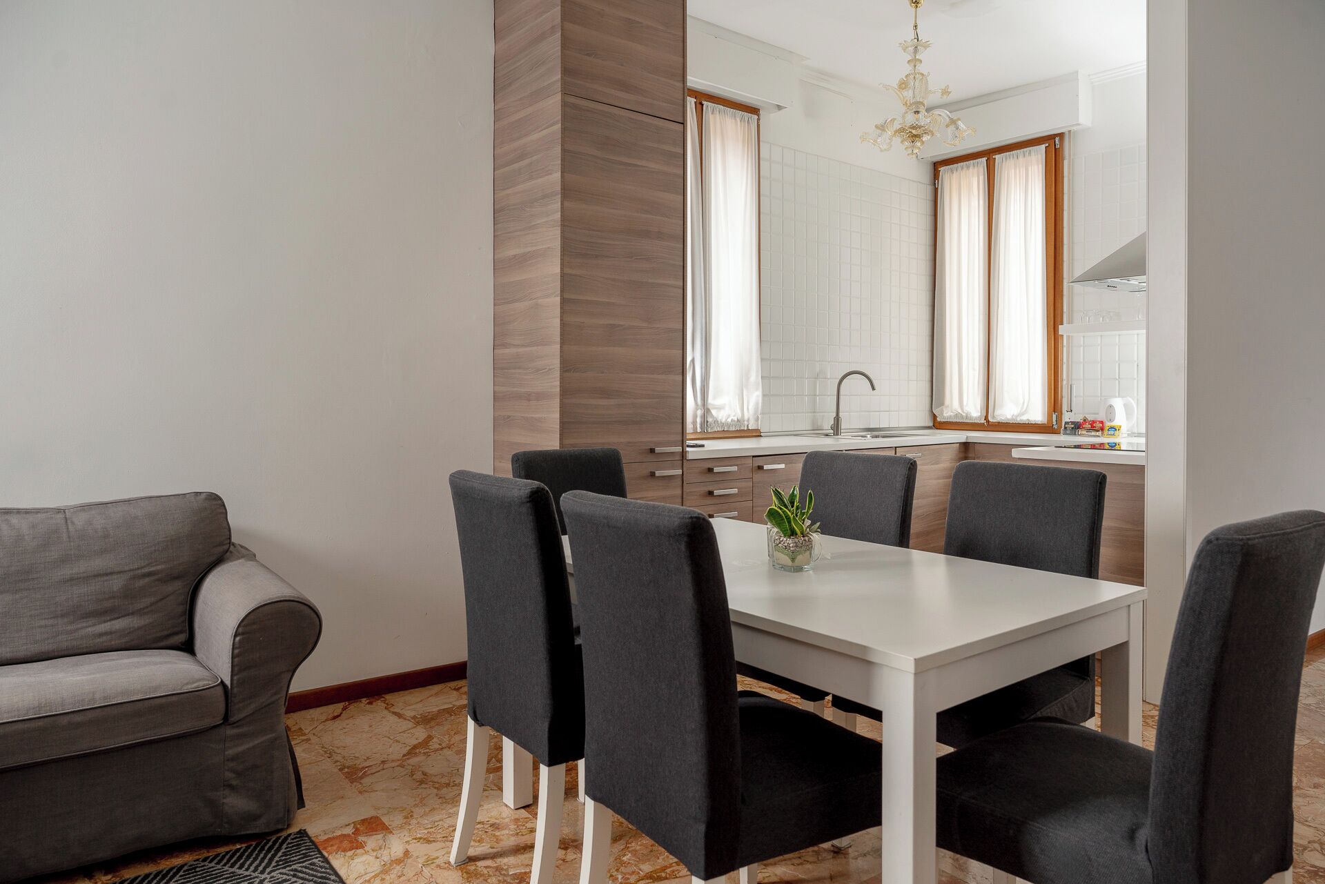 Uma área de refeições moderna com uma mesa branca e quatro cadeiras, um sofá cinza e uma cozinha ao fundo.