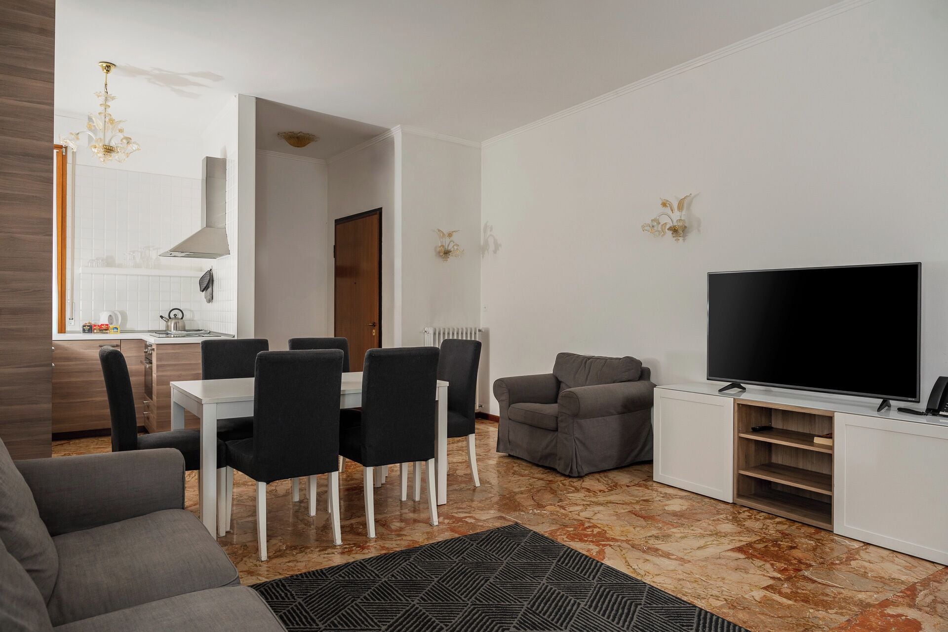 Una sala de estar moderna con televisor de pantalla plana, sofá gris, comedor con sillas y una cocina al fondo.
