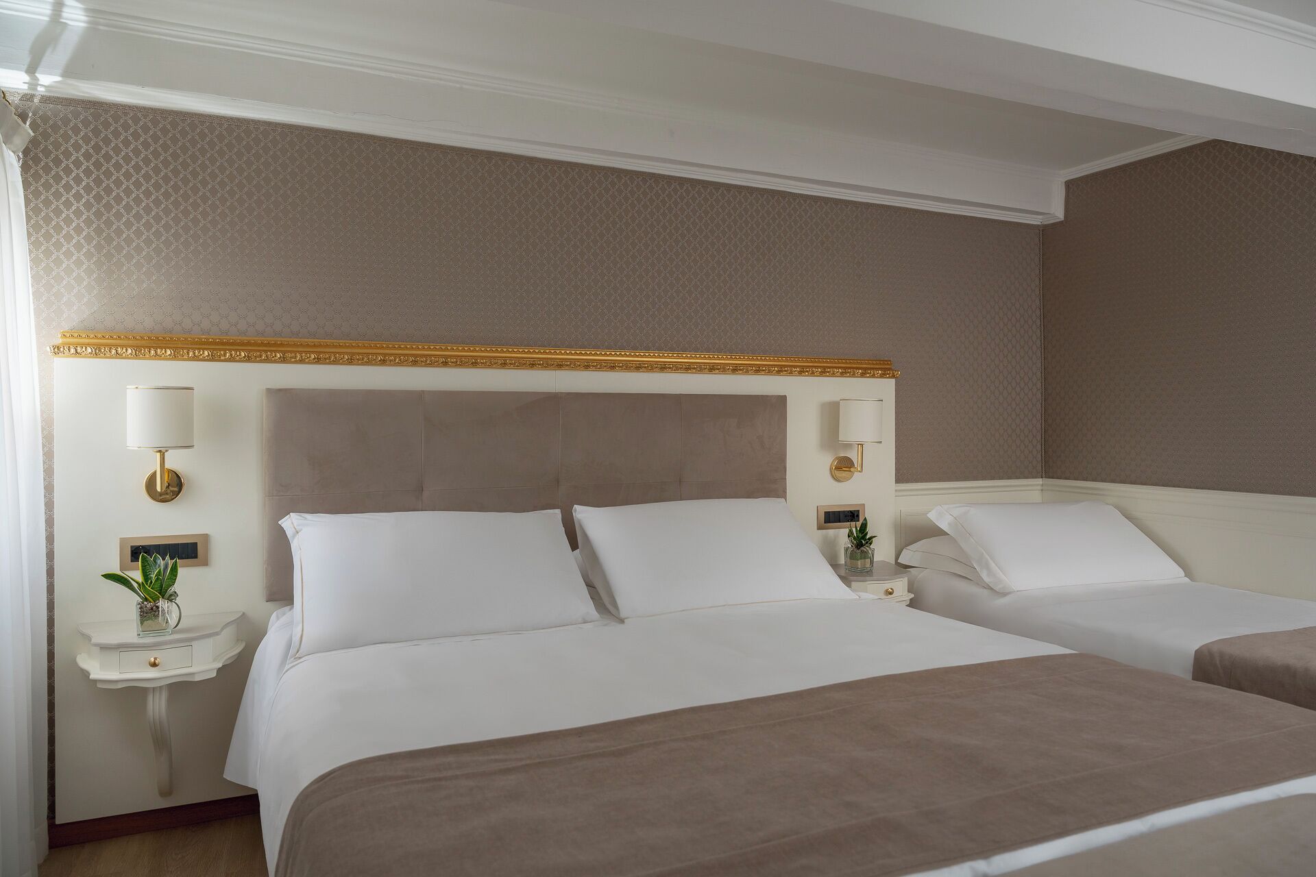 A hotel room with two beds, a large headboard, wall-mounted lamps, and a small bedside table with a plant.