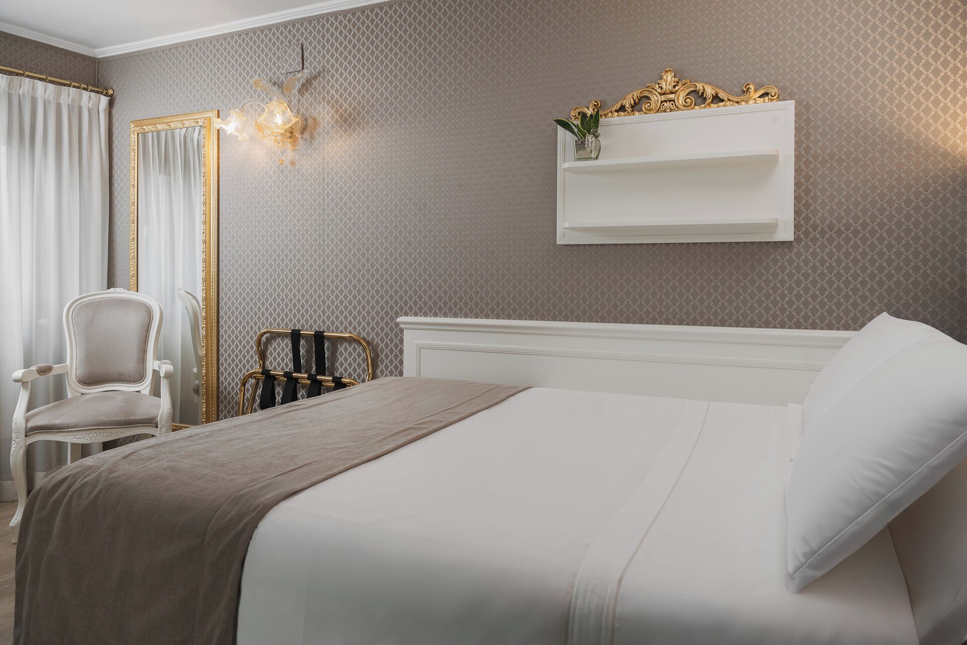 A hotel room with a bed, a chair, a nightstand, and a wall-mounted shelf with a vase.