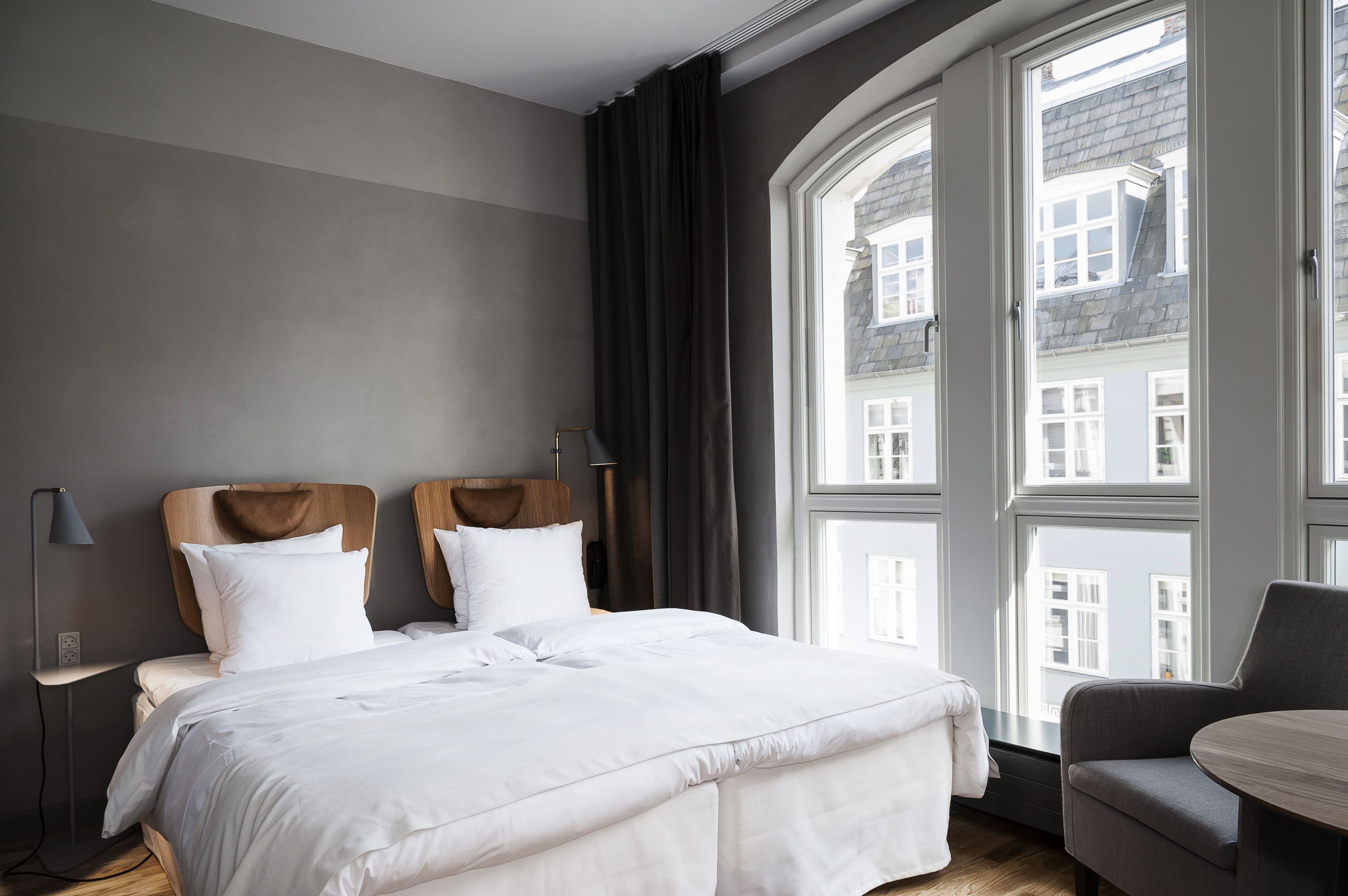 Ein Hotelzimmer mit großem Fenster, einem Bett mit weißer Bettwäsche, einem hölzernen Kopfteil, einem Nachttisch mit Lampe und einem Sessel.