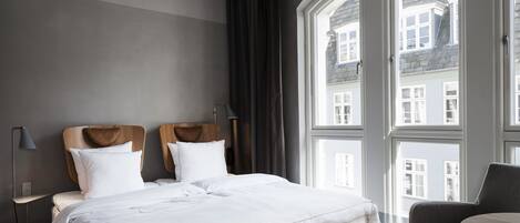 Ein Hotelzimmer mit großem Fenster, einem Bett mit weißer Bettwäsche, einem hölzernen Kopfteil, einem Nachttisch mit Lampe und einem Sessel.