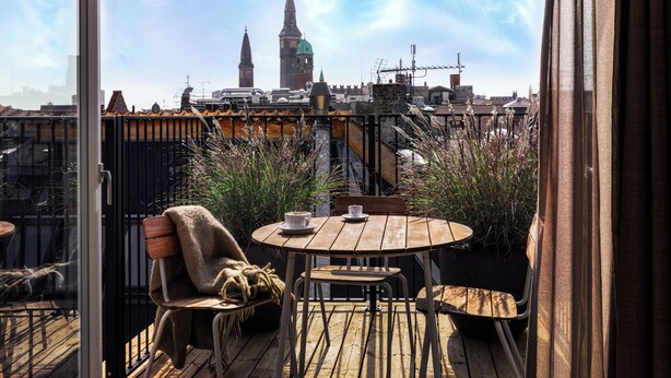 Un balcon avec une table et des chaises en bois, une vue sur le paysage urbain, notamment une tour de l’horloge, et un aperçu de la silhouette de la ville.