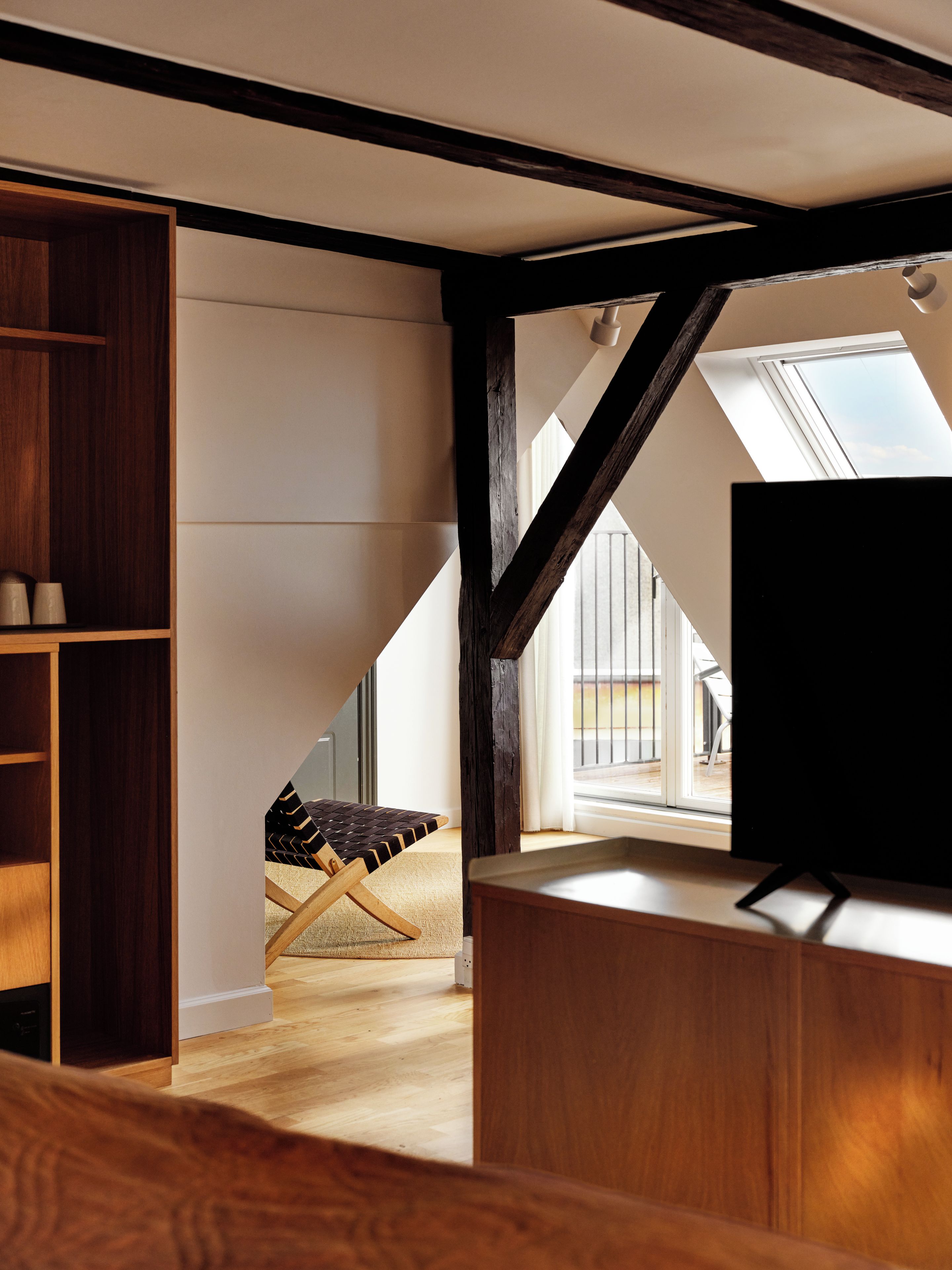 Un dormitorio estilo loft con vigas de madera a la vista, una estantería empotrada de madera, un banco acolchado y un televisor sobre un pedestal de madera.