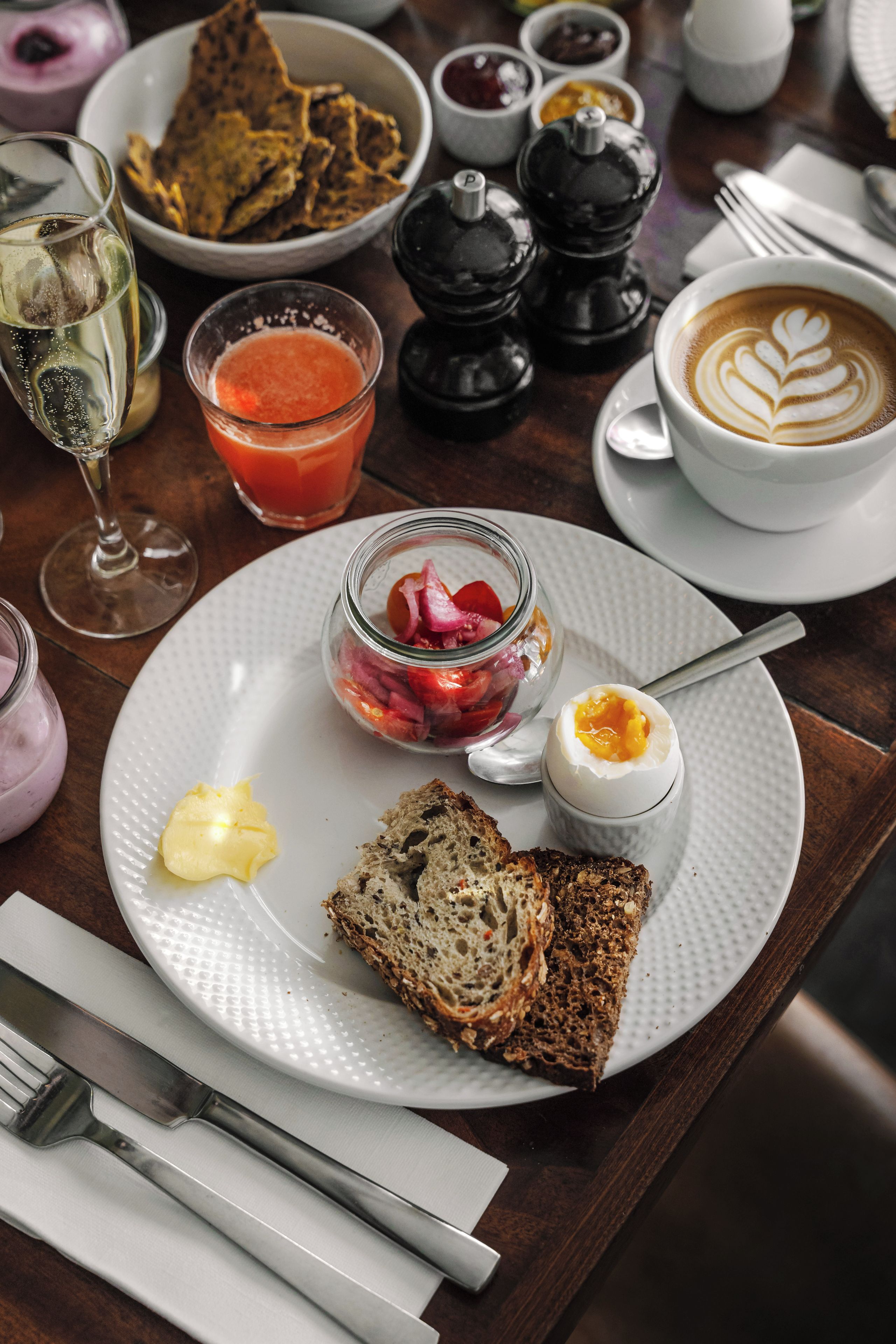 A breakfast setting with a plate of toast, a glass of orange juice, a jar of jam, a boiled egg, and a cup of coffee with latte art.