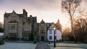Edifici de pedra històric amb arquitectura tradicional de tipus Tudor, que presenta una entrada central, un edifici de color blanc amb estructura de fusta i un jardí circumdant amb un camí de pedra.