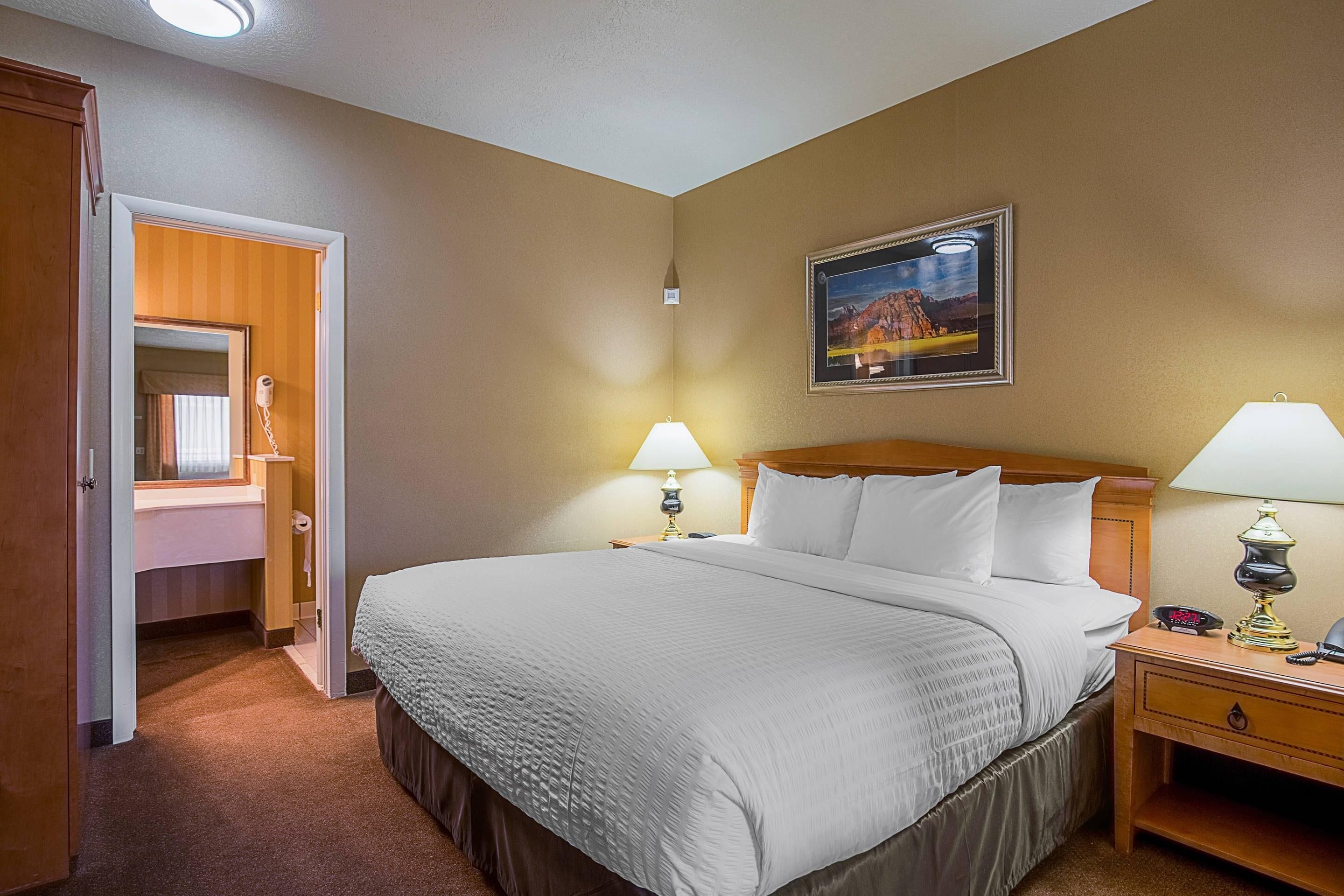 Een hotelkamer met een groot bed, twee bedlampjes, een nachtkastje, een houten hoofdbord, een ingelijst schilderij aan de muur, en een open deur naar een badkamer.
