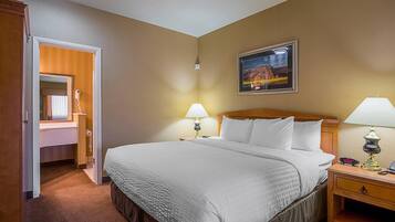 Quarto de hotel com uma cama grande, duas luminárias de cabeceira, uma mesinha de cabeceira, uma cabeceira de madeira, um quadro pendurado na parede e uma porta aberta para um banheiro.
