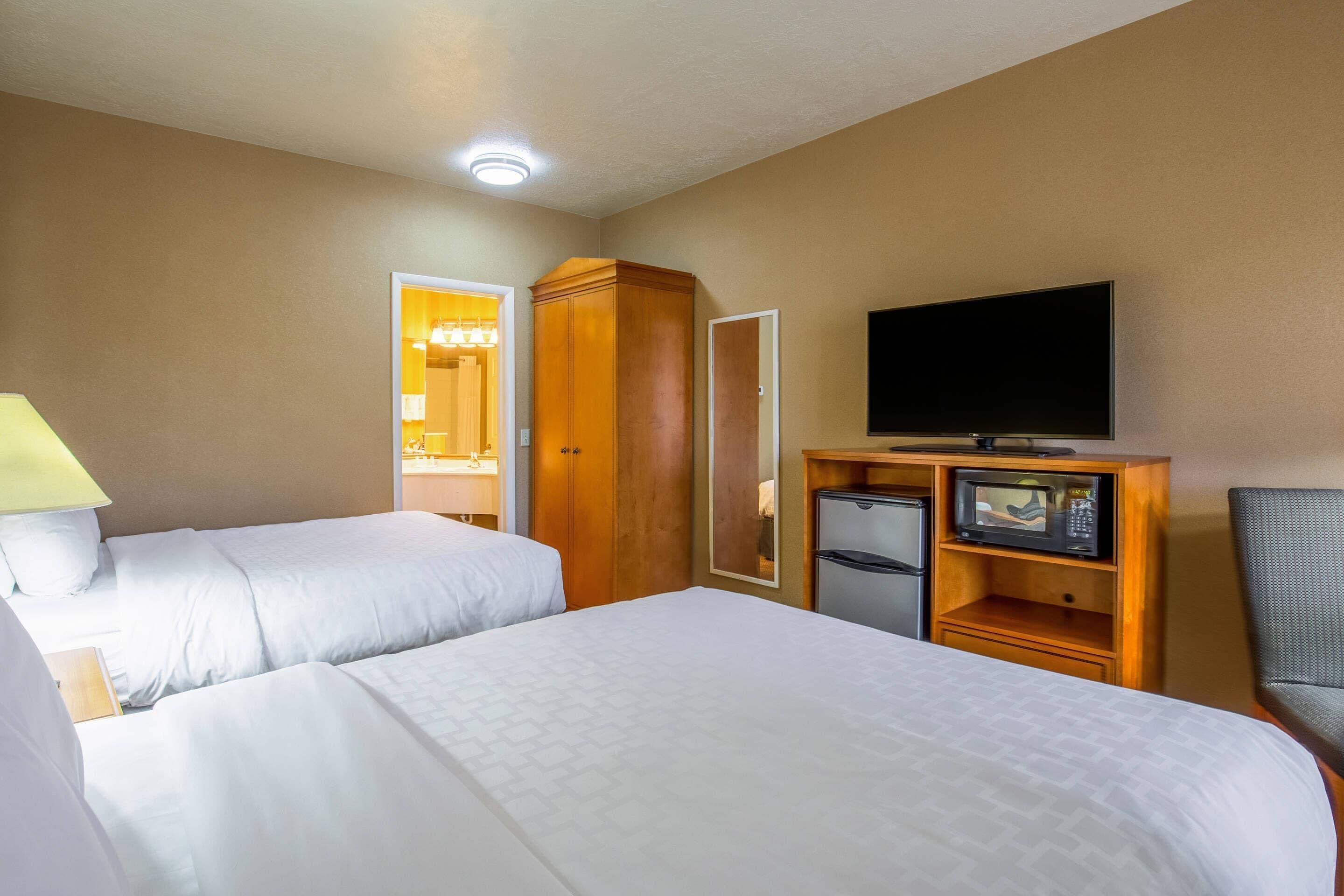 A hotel room with two beds, a television, a refrigerator, and a wooden wardrobe.