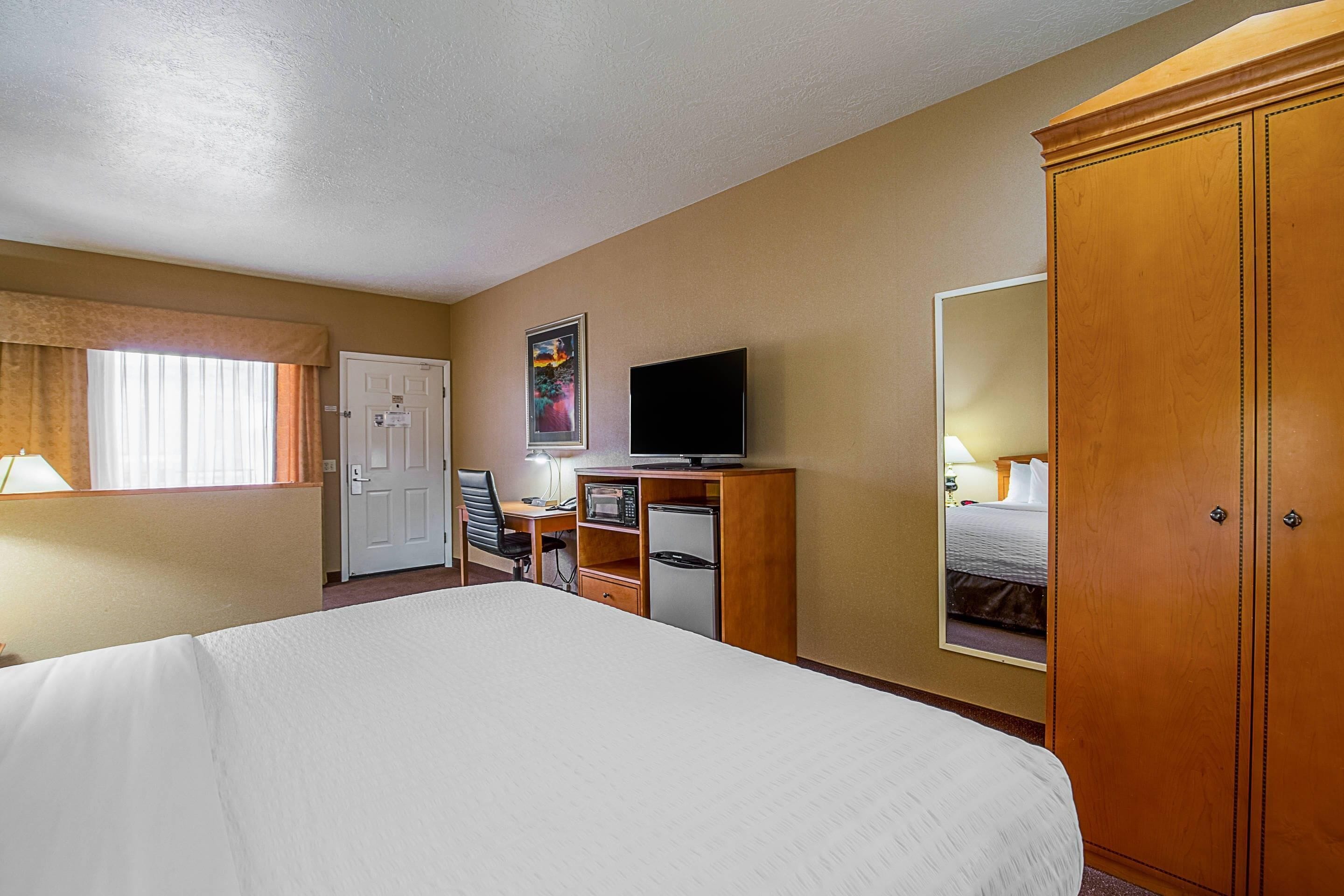 Una habitación de hotel con una cama grande, un escritorio con computadora, televisión, refrigerador y armario.