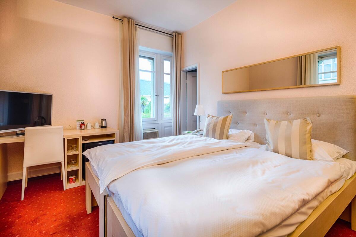 A hotel room with a bed, a desk with a television, a mirror, and a window with curtains.