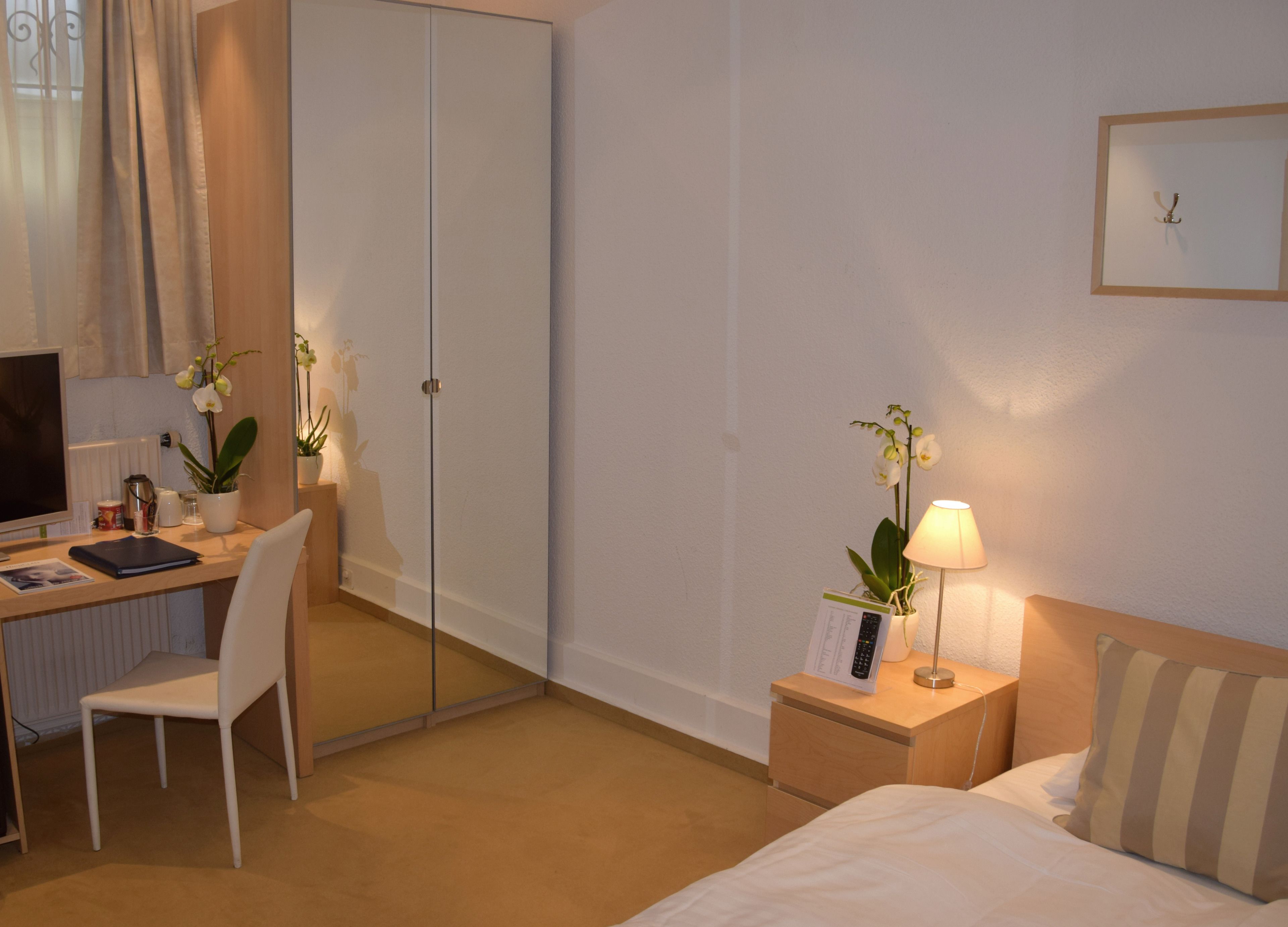 A hotel room with a bed, a desk with a computer, a chair, a bedside table with a lamp, and two potted orchids.