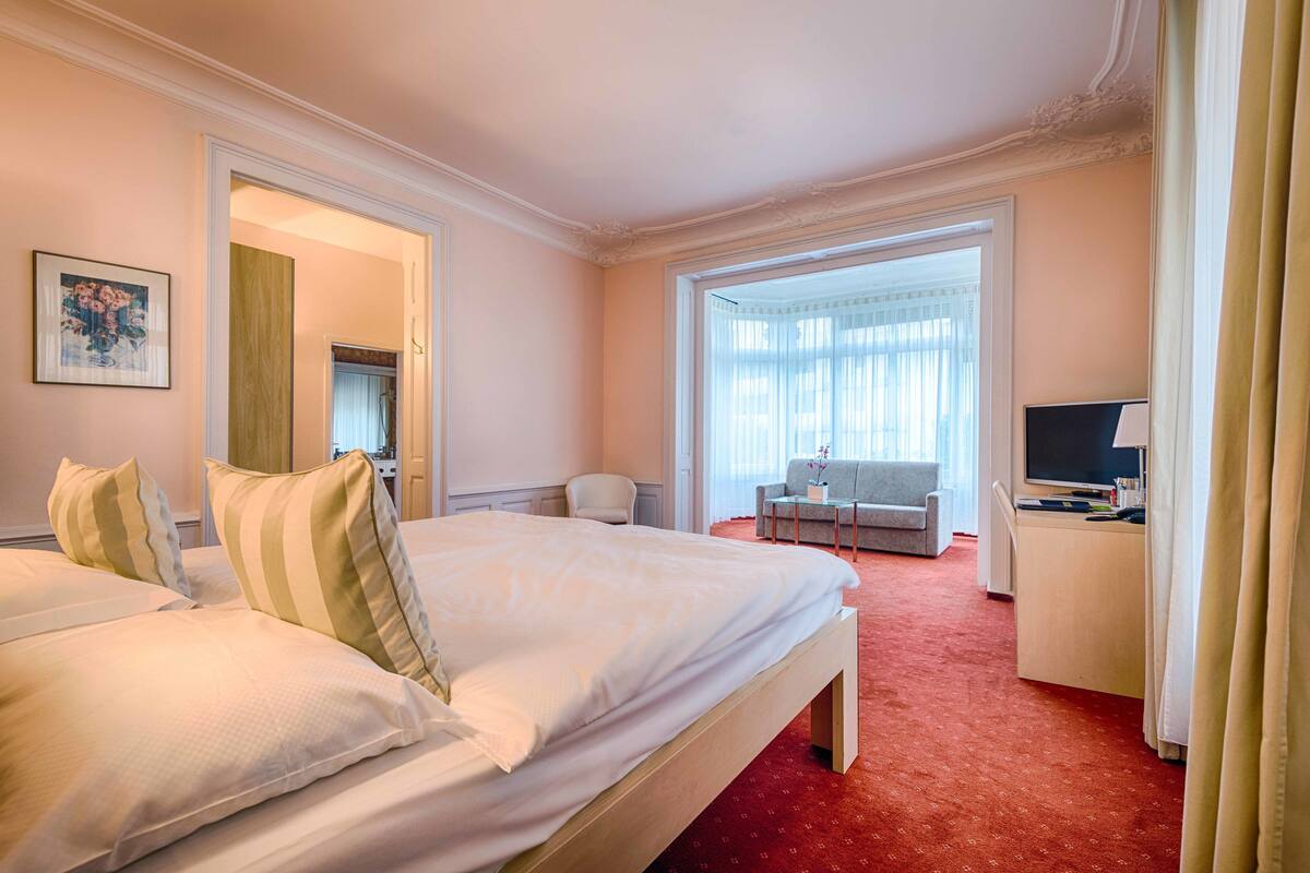 A hotel room with a large bed, a desk with a computer, a chair, and a window with curtains.