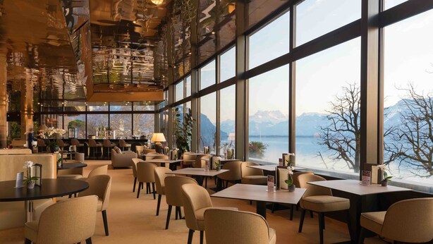Een ruime eetruimte met grote ramen die uitzicht bieden op bergen en een meer. Tafels en stoelen zijn klaar voor het dineren.