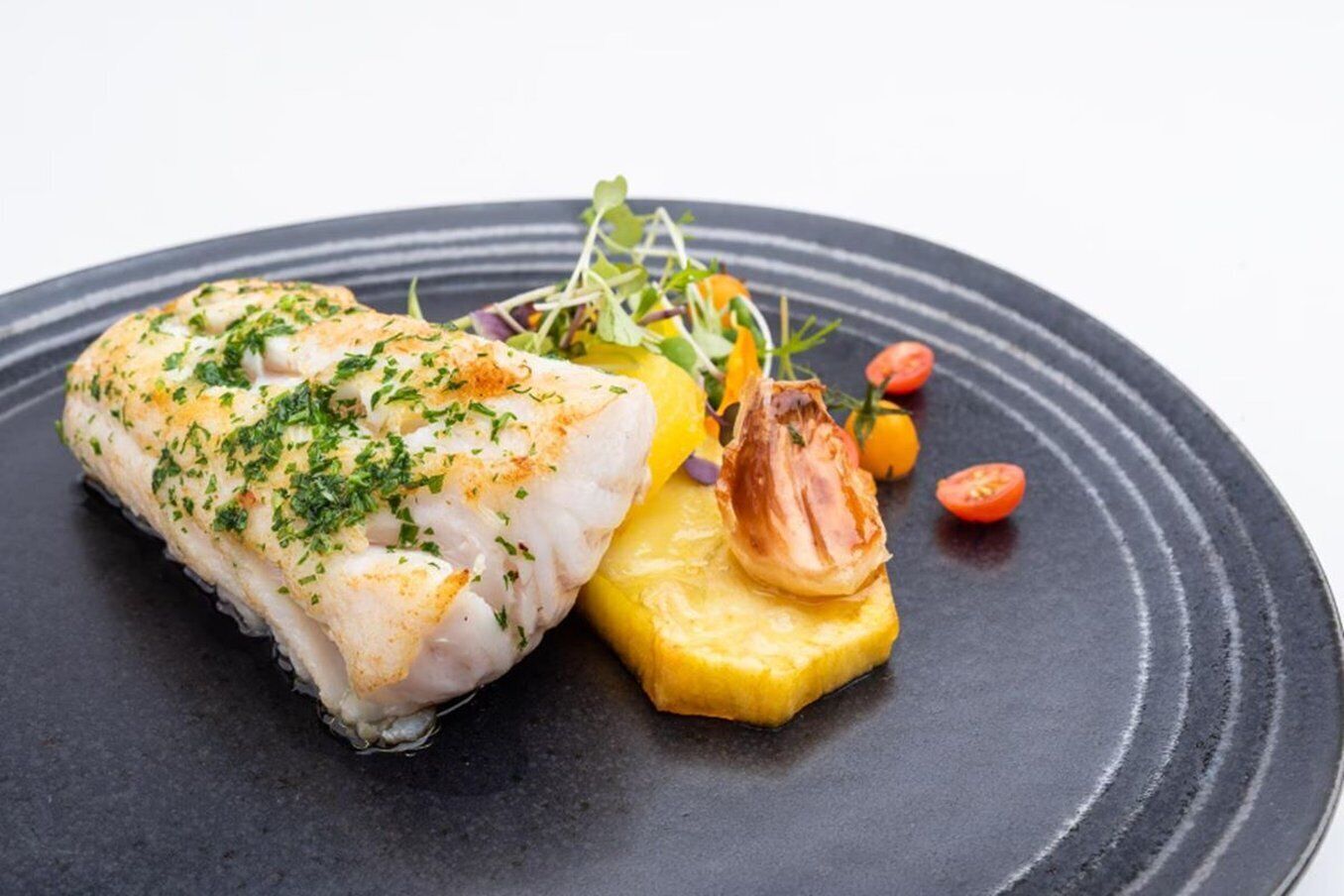 Un plato con un trozo de pescado, una rodaja de verdura amarilla y algunas guarniciones.