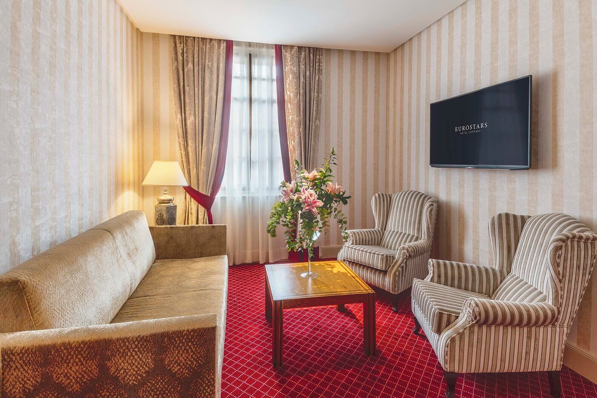 Une chambre d’hôtel avec un canapé, deux fauteuils à motifs rayés, une petite table sur laquelle est posé un vase de fleurs, une télévision et un papier peint à rayures.