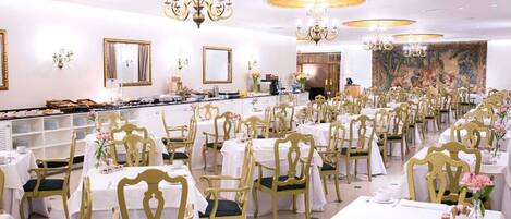 A banquet hall with rows of tables and chairs, chandeliers, and a buffet area.