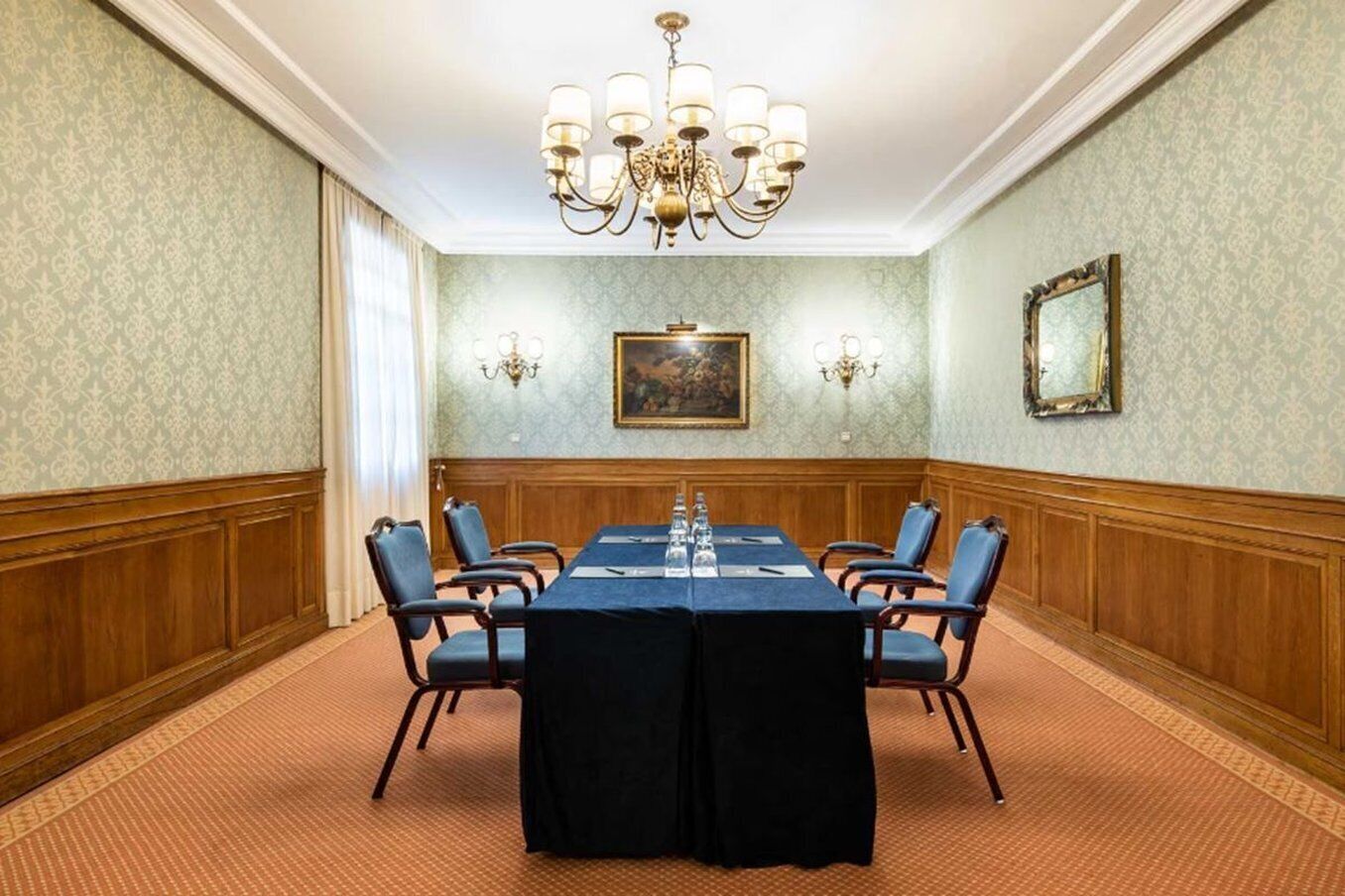 A conference room with a long table, six chairs, a chandelier, and a painting on the wall.