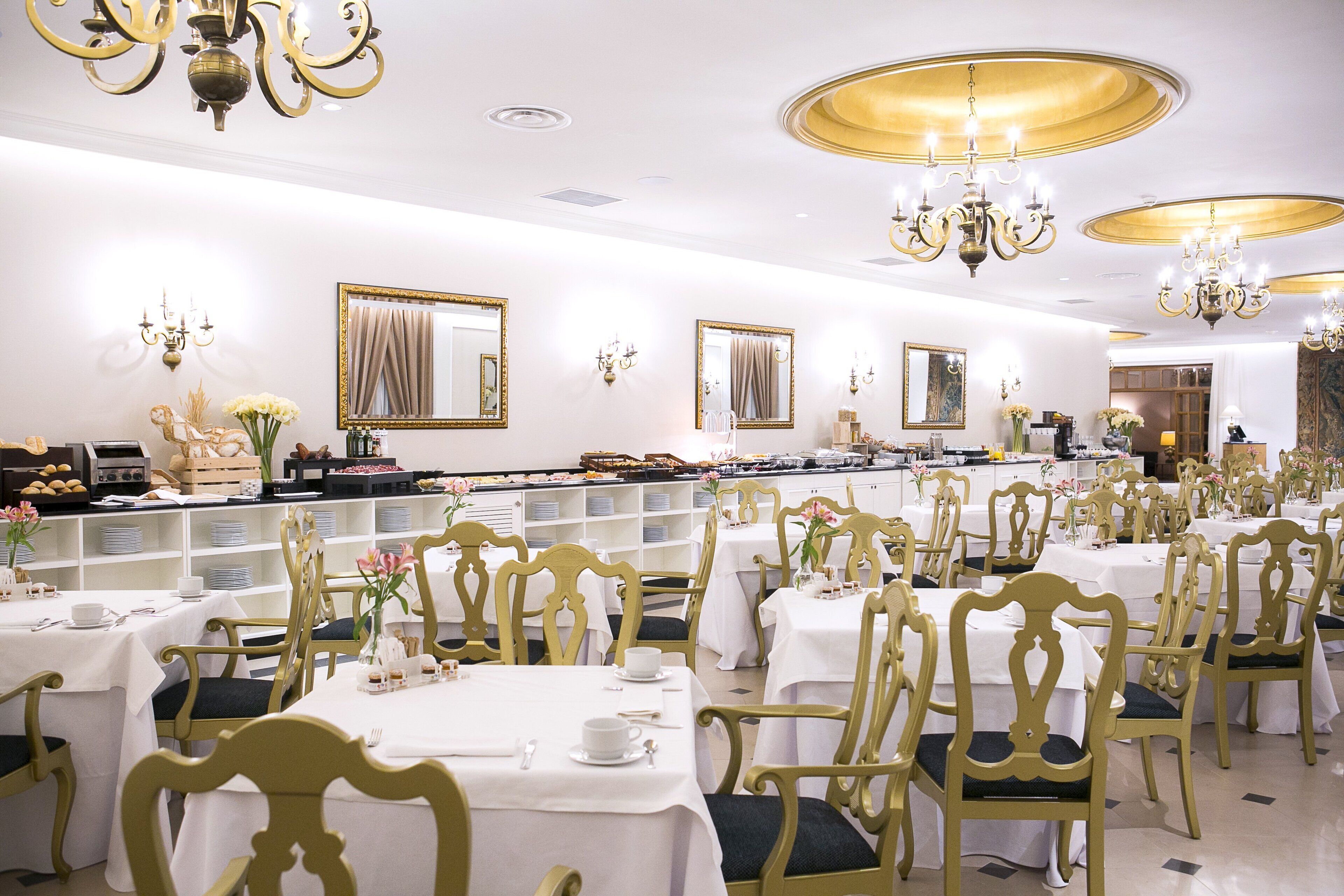 A formal dining room with multiple tables and chairs, elegant chandeliers, and decorative mirrors.