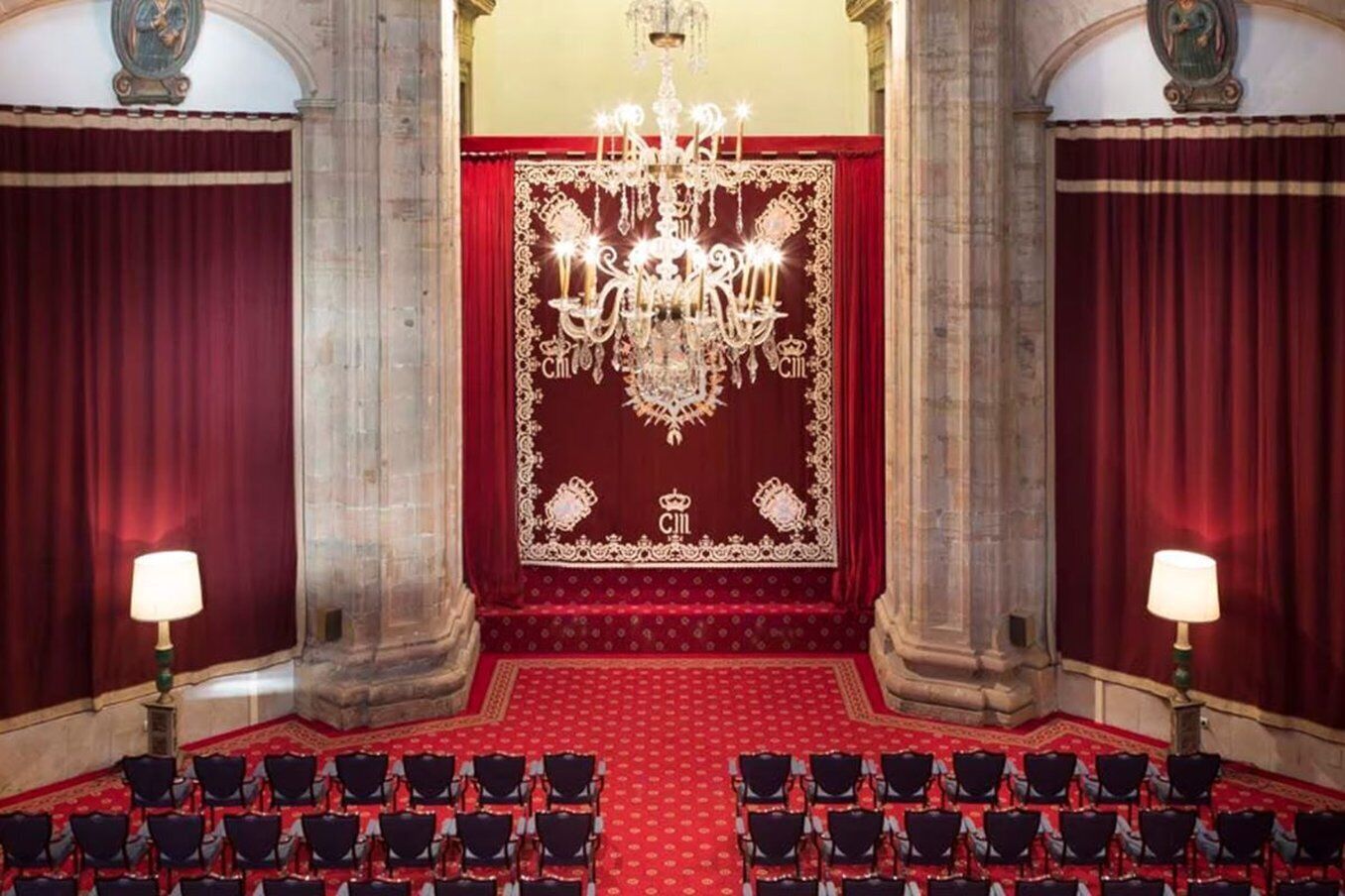 Een statige zaal met een rood tapijt, rijen stoelen, een kroonluchter en rijke gordijnen.