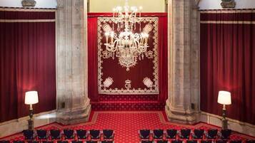 Een statige zaal met een rood tapijt, rijen stoelen, een kroonluchter en rijke gordijnen.