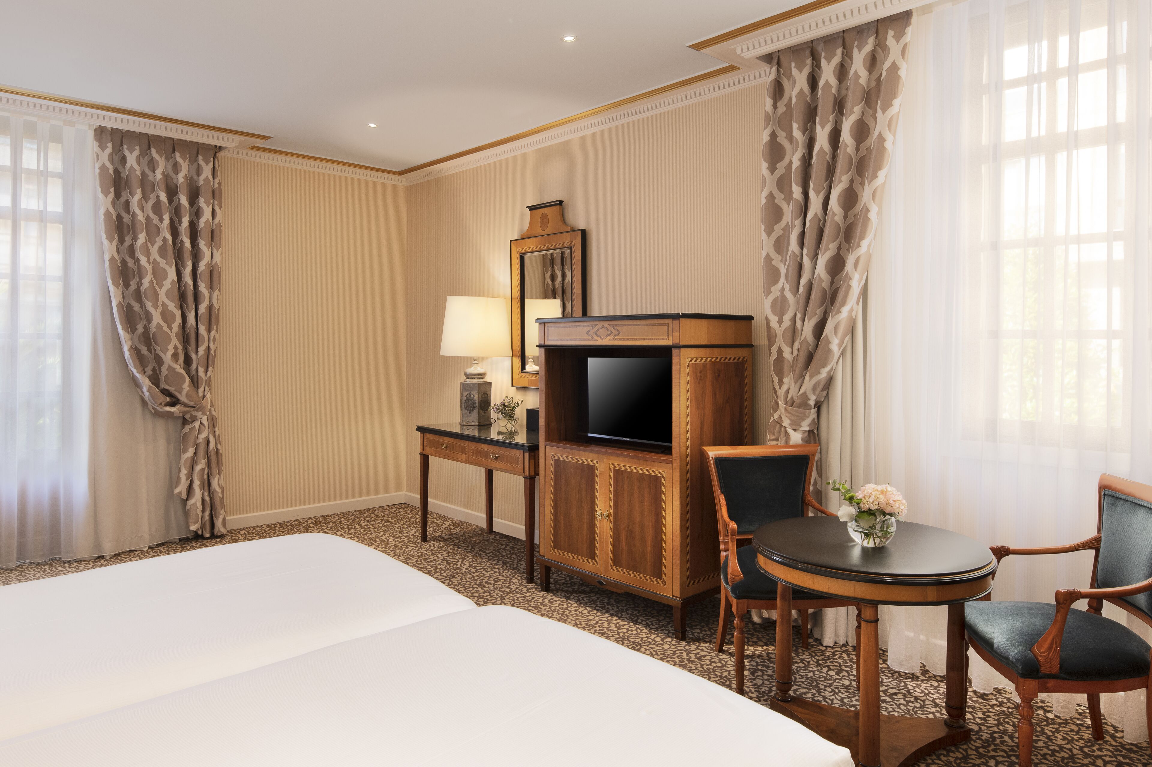 A hotel room with a bed, a desk with a television, a chair, and a small table with a vase of flowers.