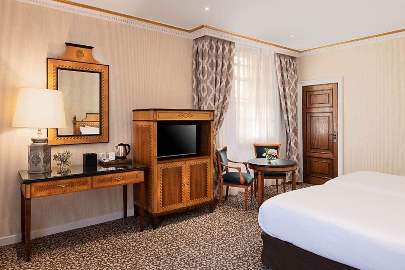 A hotel room with a bed, a desk with a television, a chair, a small table with a vase, and a door.