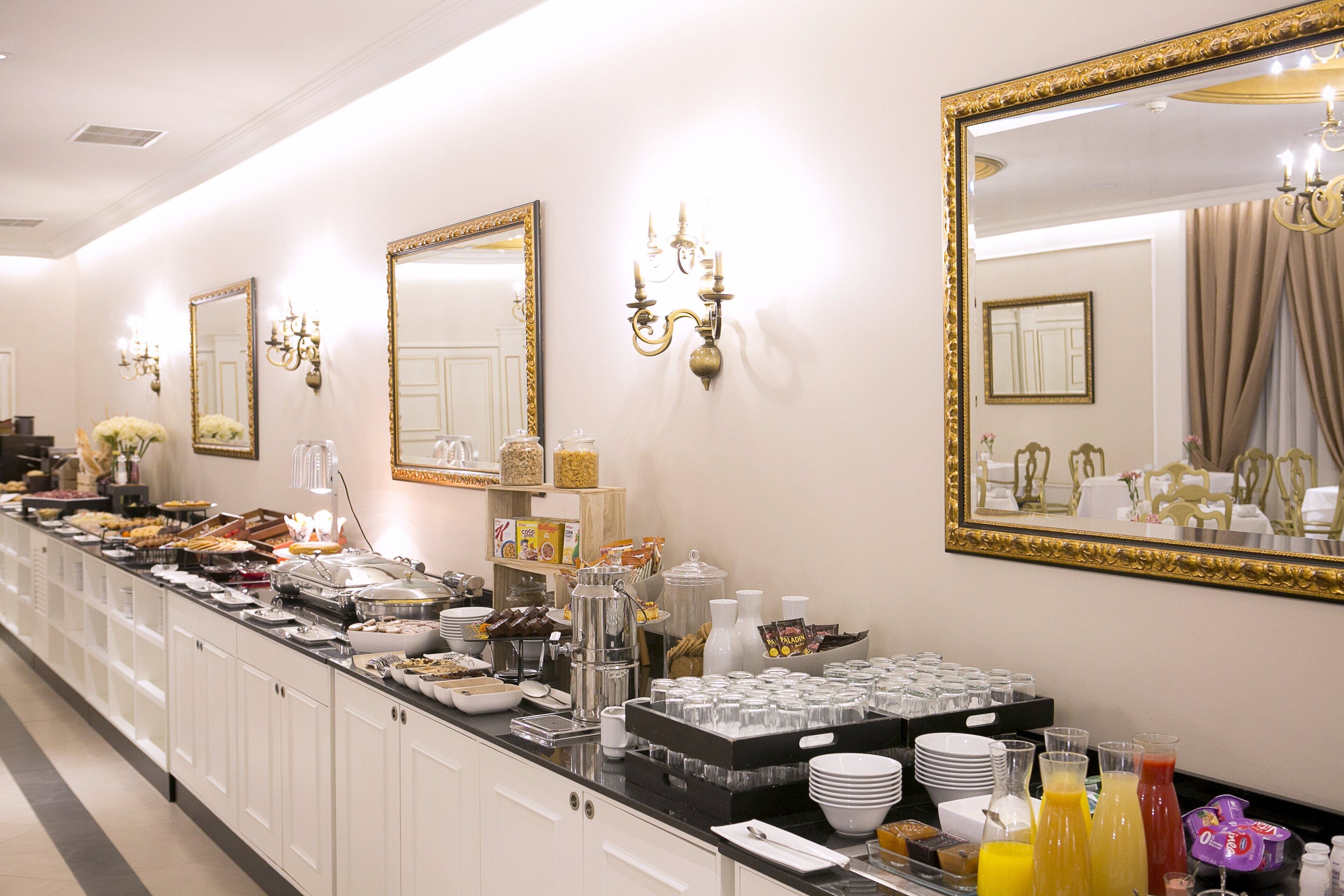 A buffet setup with various food items and beverages, a mirror, and wall-mounted light fixtures.