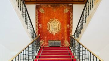 Una gran escalera con peldaños alfombrados de rojo y barandillas negras, que conduce a un salón con un tapiz rojo ornamentado de gran tamaño que presenta un escudo de armas.