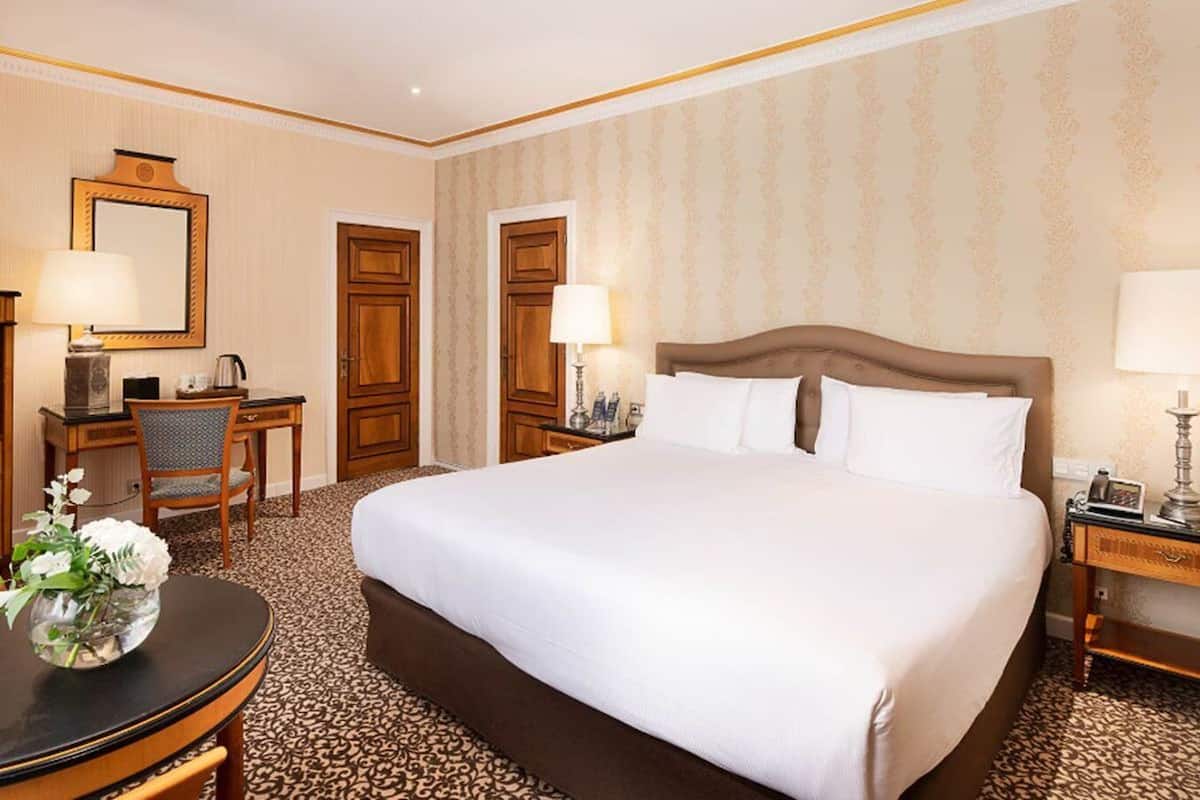 Ein Hotelzimmer mit einem großen Bett, einem Schreibtisch mit Stuhl, einem kleinen Tisch mit Blumenvase und zwei Nachttischlampen.