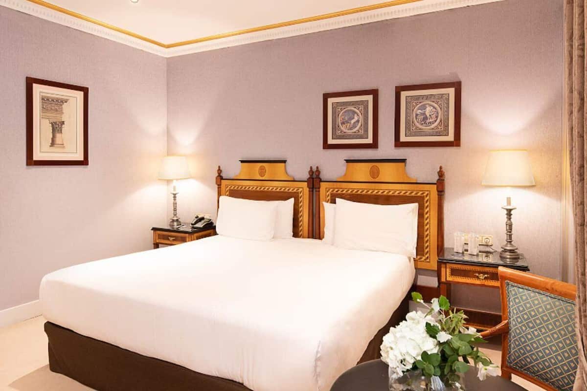 Ein Hotelzimmer mit einem großen Bett, zwei Nachttischen, einem Beistelltisch, einem Sessel und einer Vase mit Blumen.