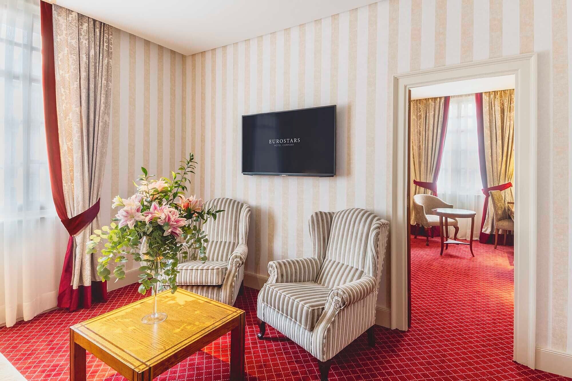 A hotel room with striped wallpaper, a flat-screen TV, two striped armchairs, a small table with a vase of flowers, and a red carpeted floor.